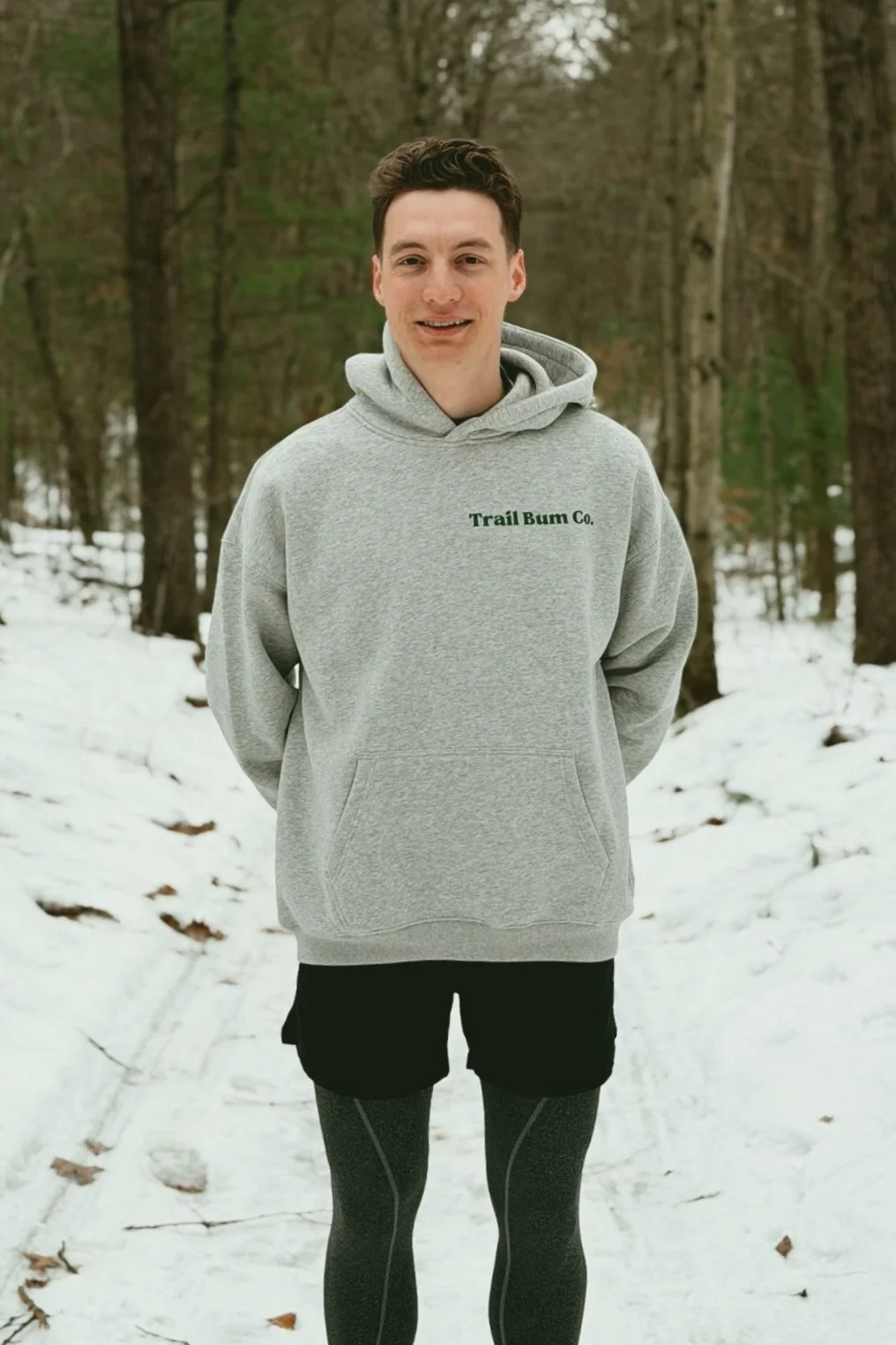 A young man smiling in a snowy forest, wearing a gray hoodie with 'Trail Bum Co.' printed on it, black shorts, and black running leggings.