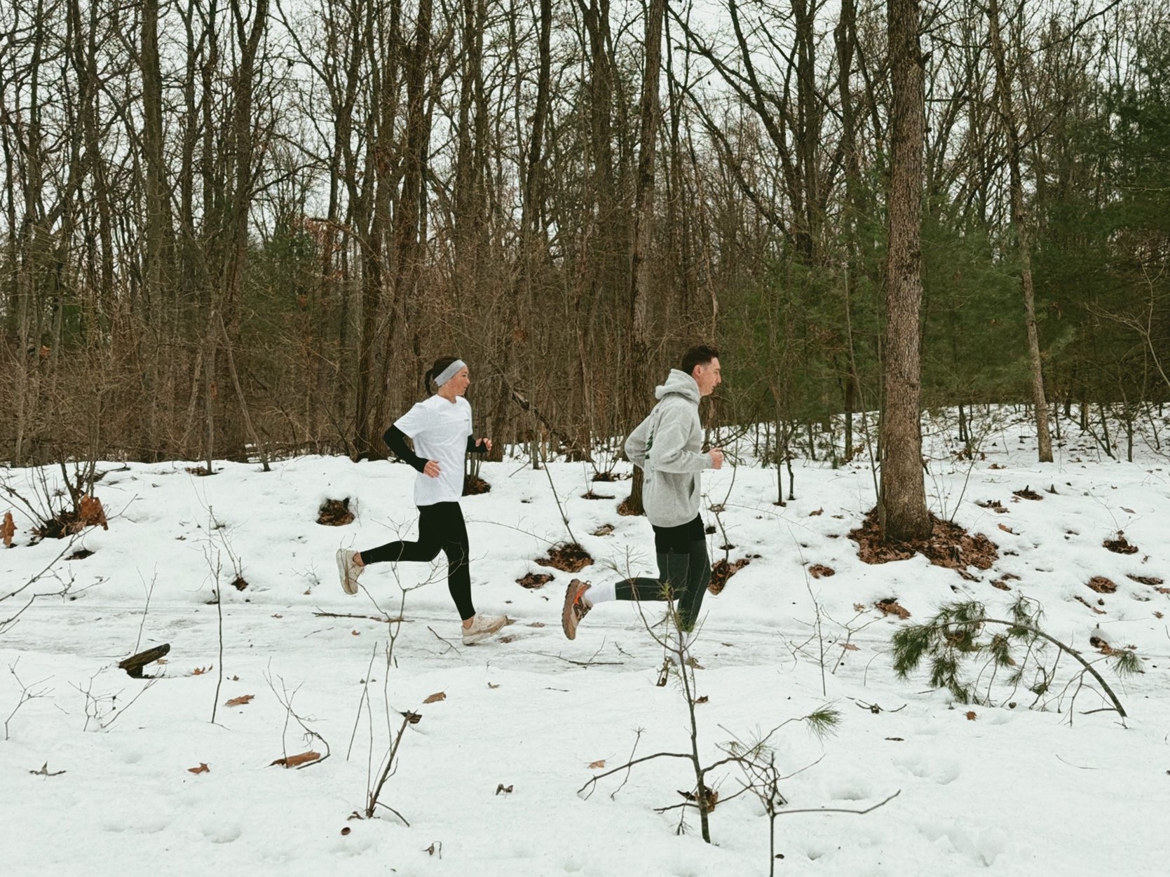 Two people running in a snowy forest, one woman and one man, wearing athletic clothing.