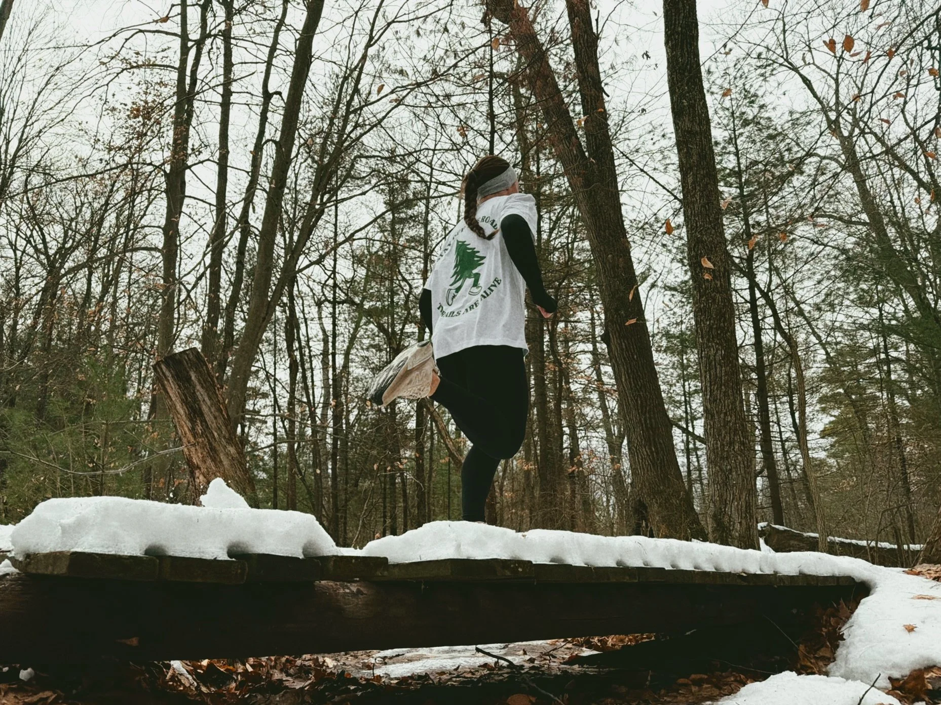 A person jogging on a snow-covered wooden bridge in a forest with tall trees and a cloudy sky.