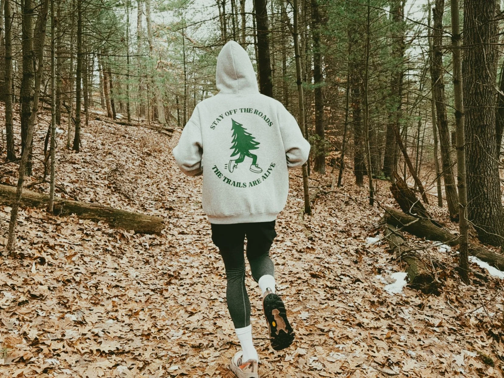 Person running through a wooded forest trail with fallen leaves on the ground. The person is wearing a gray hoodie with a green pine tree graphic and the text 'Stay off the roads, the trails are alive' on the back, black leggings, and running shoes.