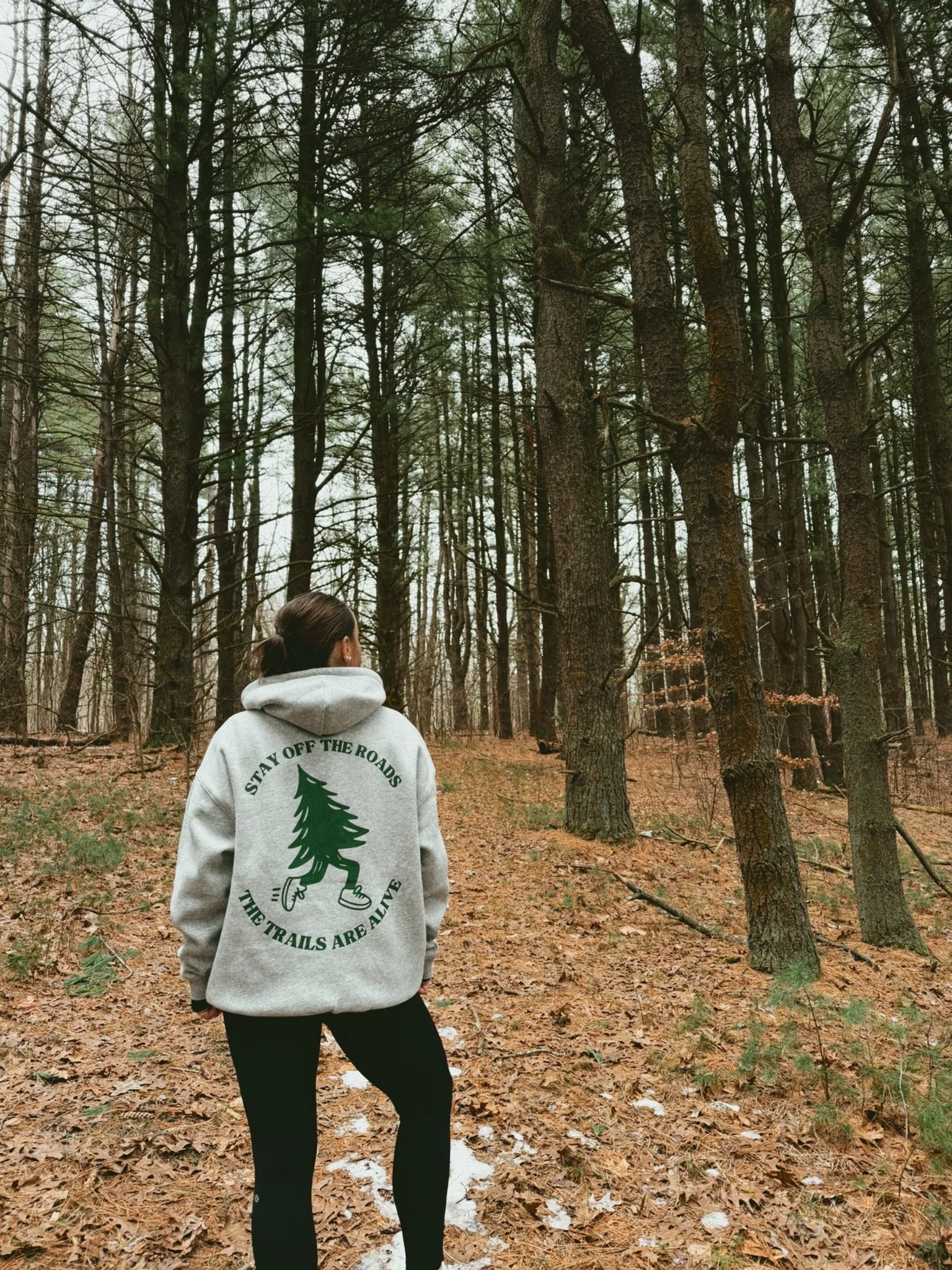 A woman stands in a wooded forest during winter wearing a gray hoodie with a green tree illustration and the words "Stay Off the Roads, The Trails Are Alive."