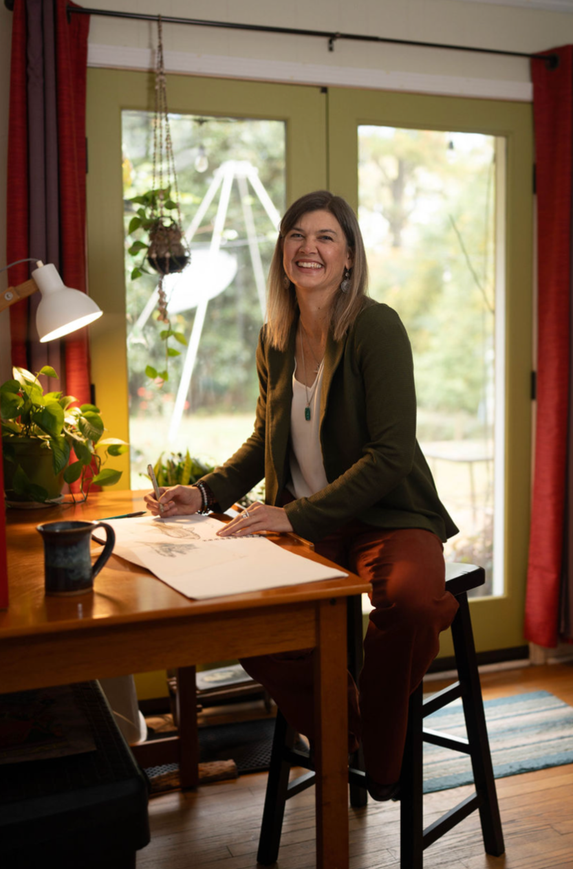 A woman with shoulder-length brown hair, wearing a dark blazer and white top, sitting at a wooden table in a cozy room, smiling as she sketches on a large sheet of paper. The room has large glass doors with green framing, red curtains, indoor plants, and a swing outside visible through the glass.