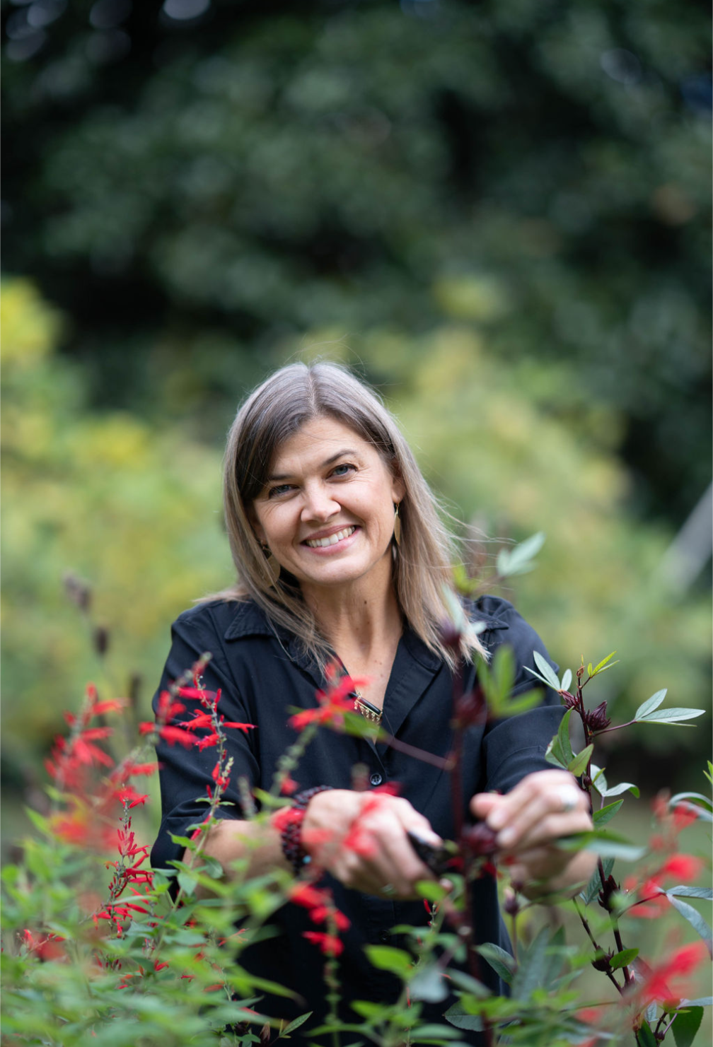 A woman with shoulder-length brown hair smiling while trimming red and green plants outdoors in a lush garden.