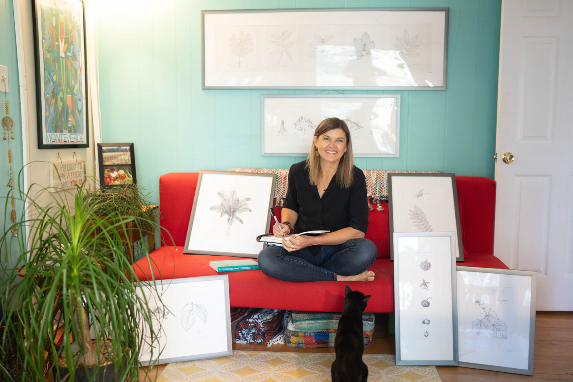 Gini Knight sitting cross-legged on a red sofa in a brightly lit room, surrounded by framed botanical illustrations, with a small black cat sitting on the floor in front of her.