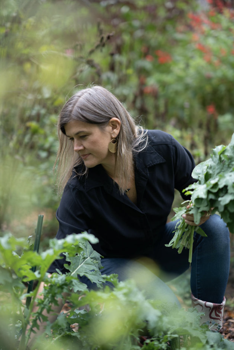 A woman crouching in a garden, harvesting leafy greens with a gentle expression surrounded by lush greenery and colorful flowers.