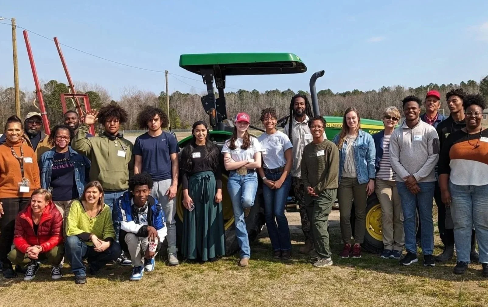 A group of people standing outdoors in front of a green John Deere tractor, with some smiling and waving, on a sunny day.
