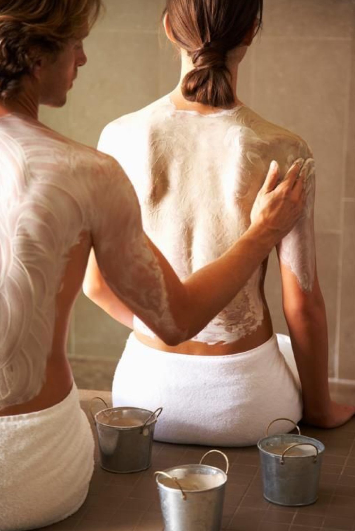 Two women in towels have a spa or bath session, applying a creamy facial or body treatment, with metallic buckets on the floor.