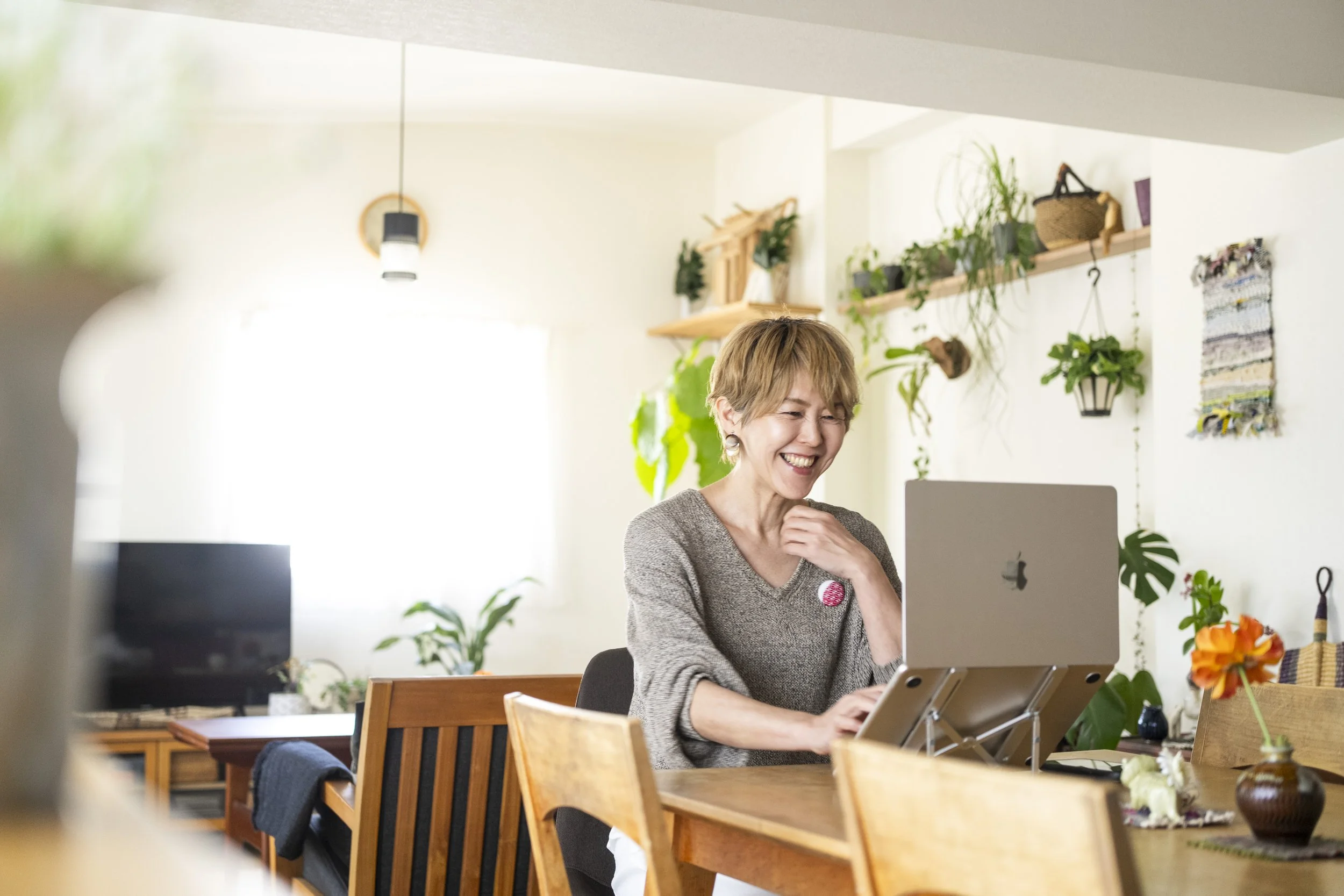 室内でコンピュータを使い笑顔の女性が作業している風景。背後には壁に掛けられた植物や装飾品。明るく温かい雰囲気。