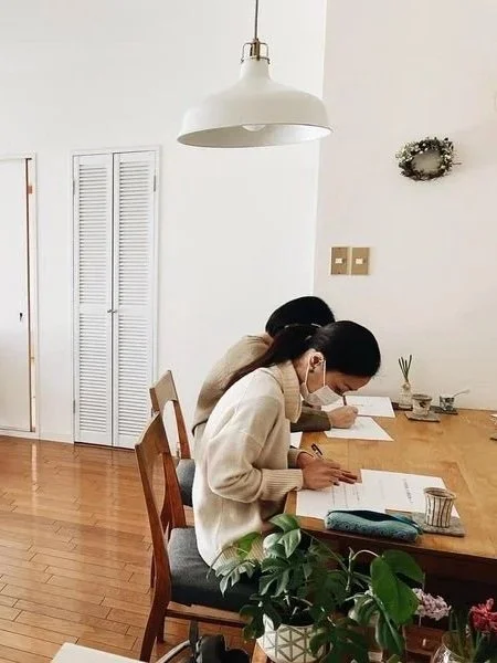 リビングルームのダイニングテーブルで3人の女性が勉強や作業をしている。白い壁と木製の床、壁にリースが飾られている。