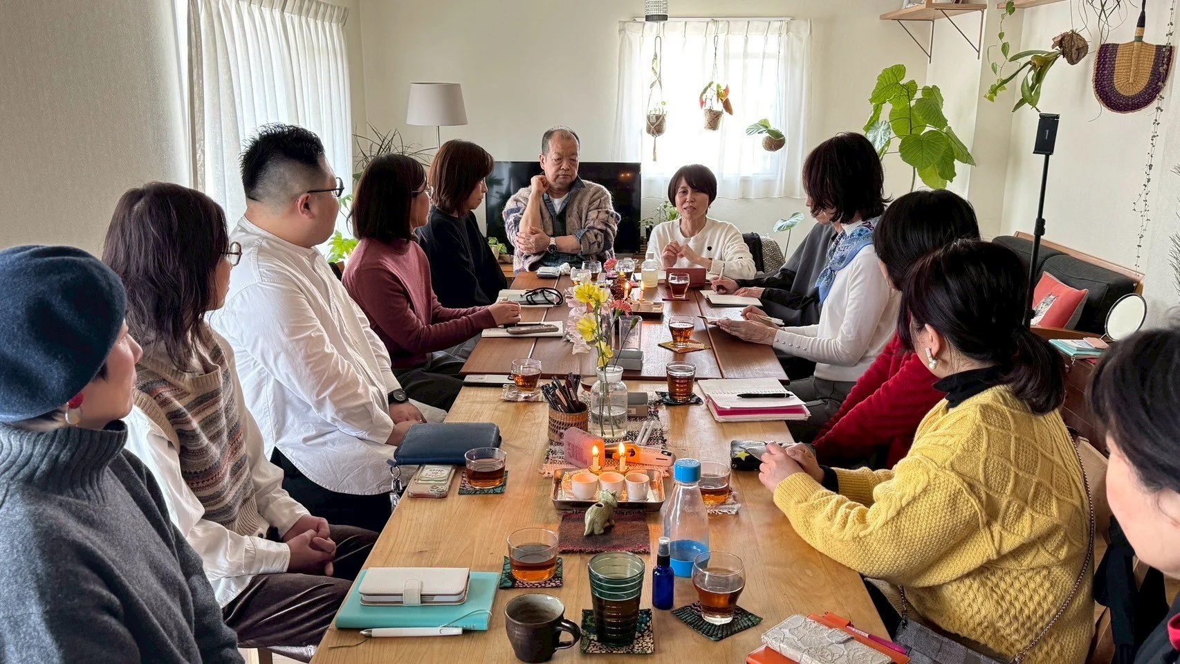 室内で会議をしている人々のグループ。長い木製のテーブルに人々が座り、多くのノートや飲み物、キャンドル、飾りが置かれている。背景には観葉植物とカーテンが見える。
