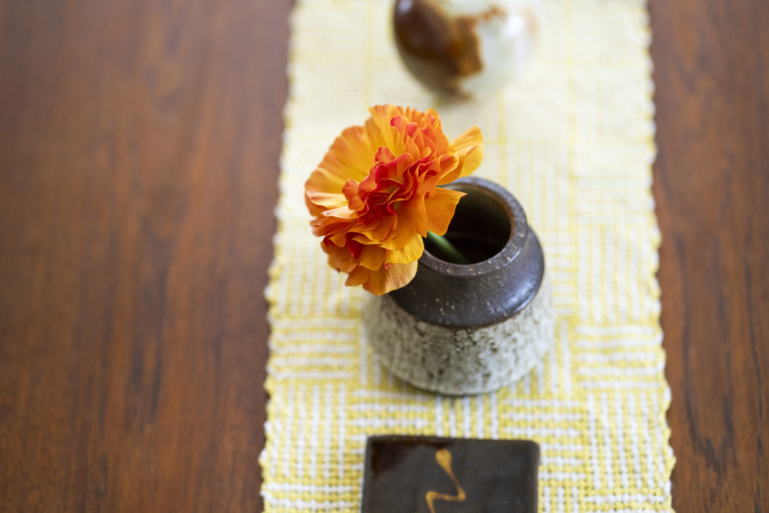 橙色のカーネーションの花が黒い花瓶に入っている。花瓶は石の上に置かれ、黄色のチェック柄の布の上に置かれている。背景は木製のテーブルとぼやけた背景。