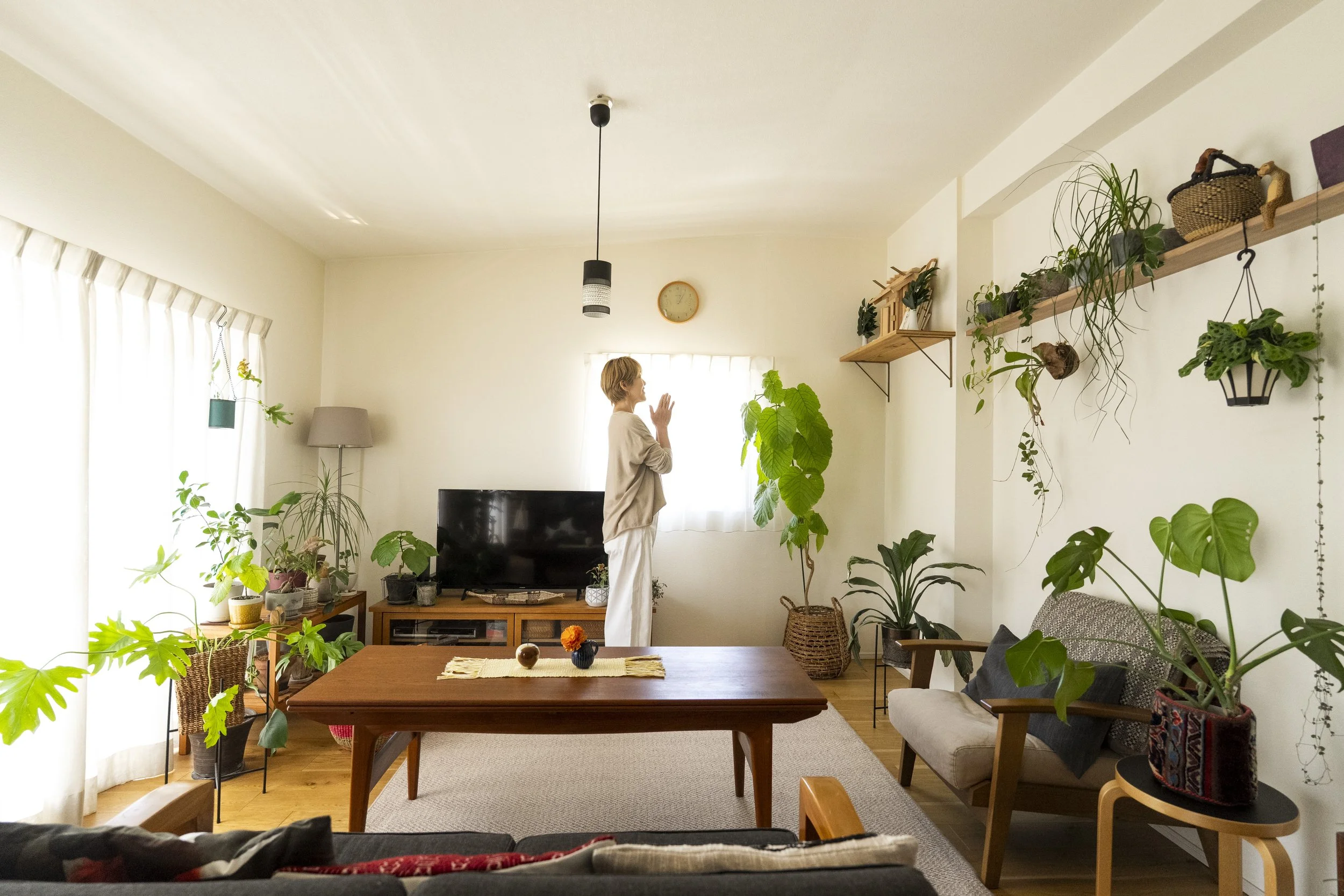 多数の観葉植物が置かれたリビングルーム。壁にかけられた時計の下には女性が立ち、手を合わせて祈る姿勢をしている。テーブルの上には花や飾りがある。窓から自然光が差し込んでいる。