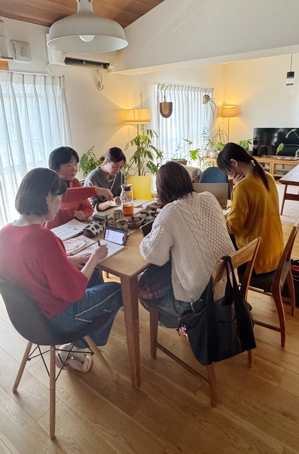 室内のリビングルームで、テーブルを囲みながら話し合っている5人の女性たち。室内には観葉植物や照明、テレビが配置されている。