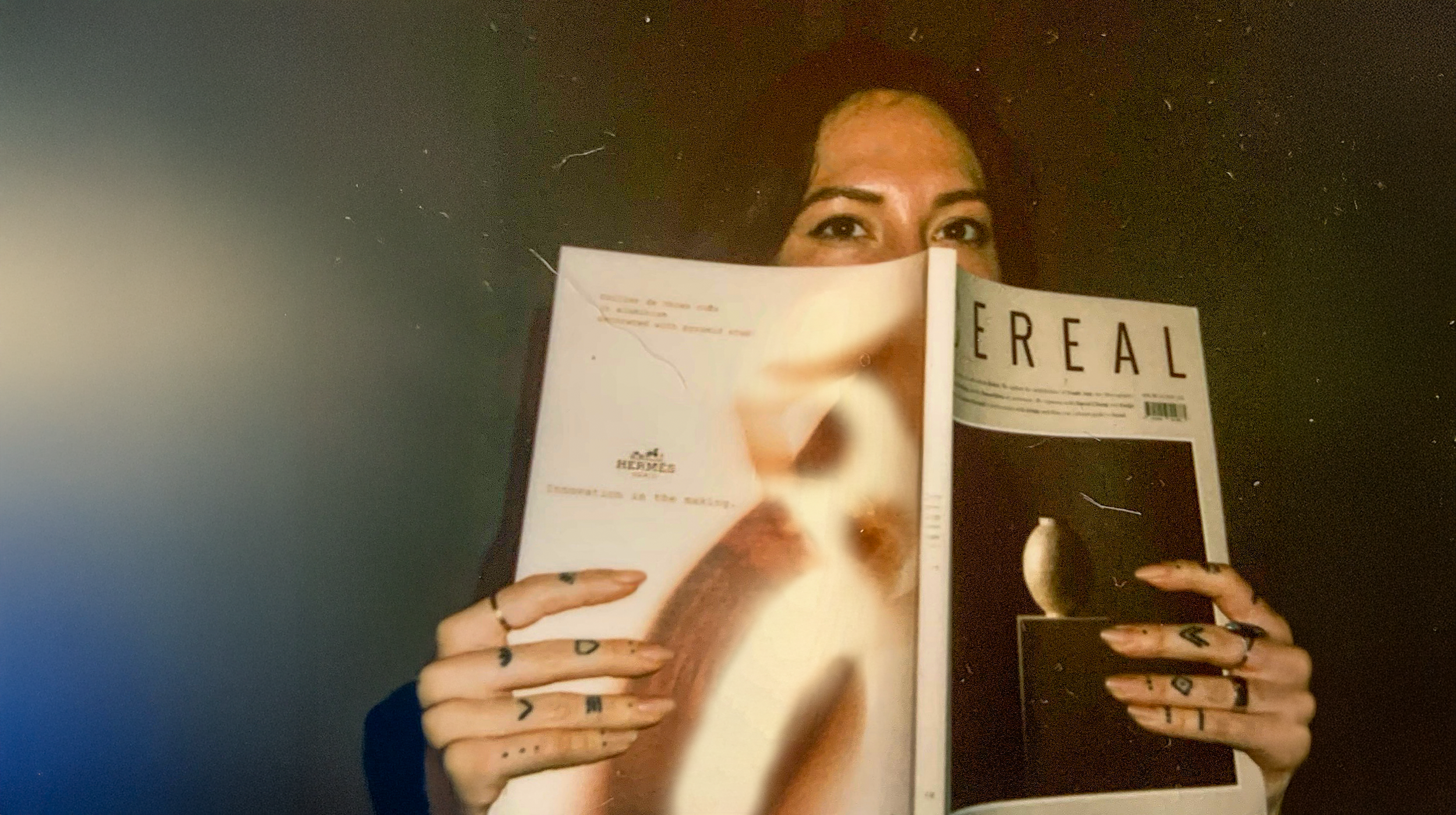 Portrait of Taylor Hammes holding an open magazine, Cereal,  that partially covers her face, revealing her eyes above the pages, in a softly lit, editorial-style photograph.