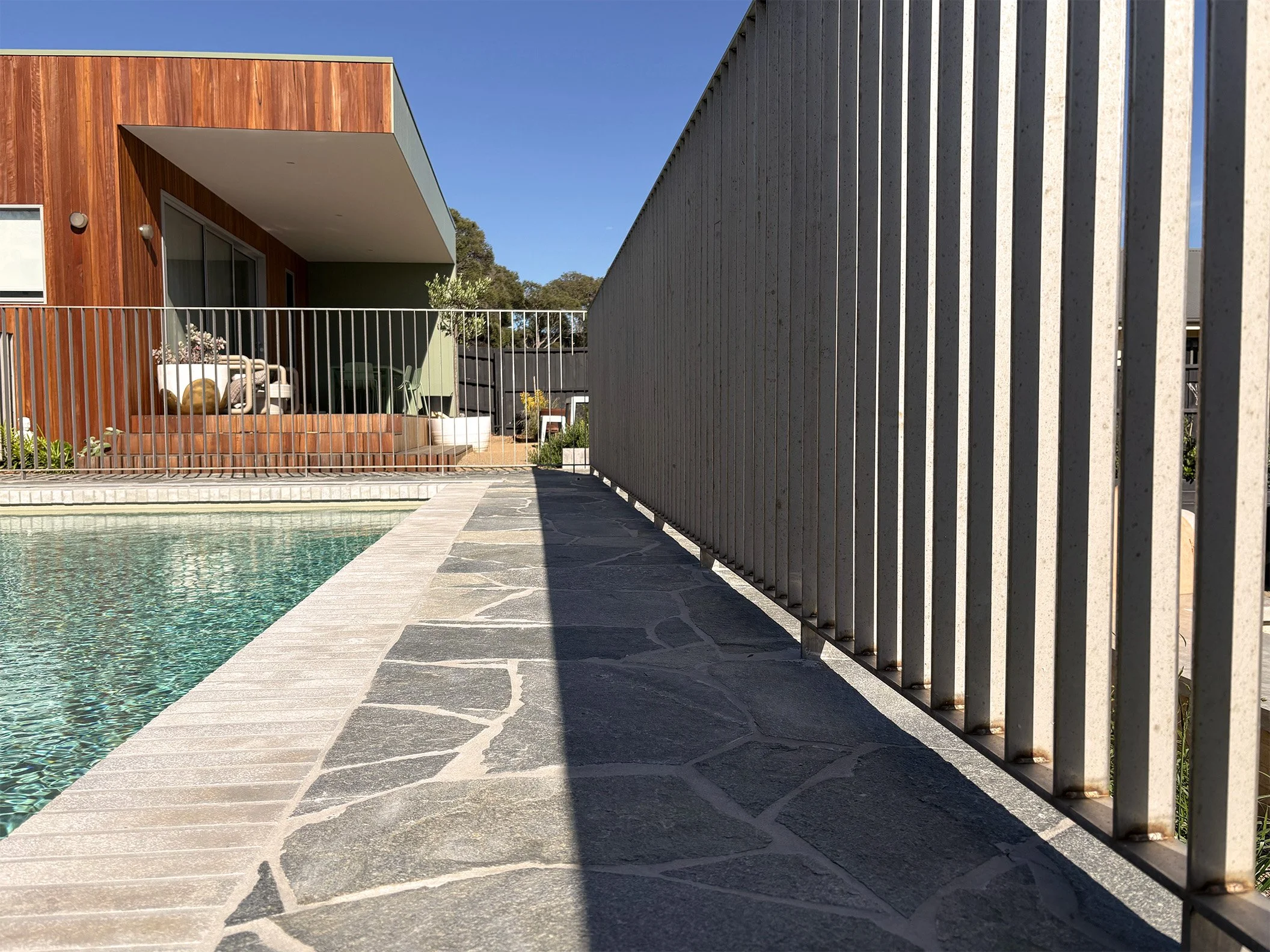 Side view of a swimming pool with a stone deck, a wooden house with a patio, and a concrete fence on a sunny day.
