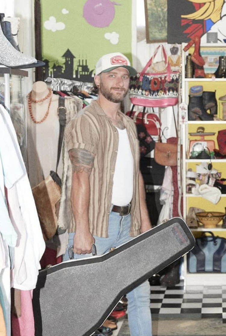 A man with a beard, wearing a striped shirt and a baseball cap, standing inside a vintage or thrift store. He is holding a guitar case, surrounded by various clothing and accessories, with colorful artwork and shelves displaying bags and shoes in the background.