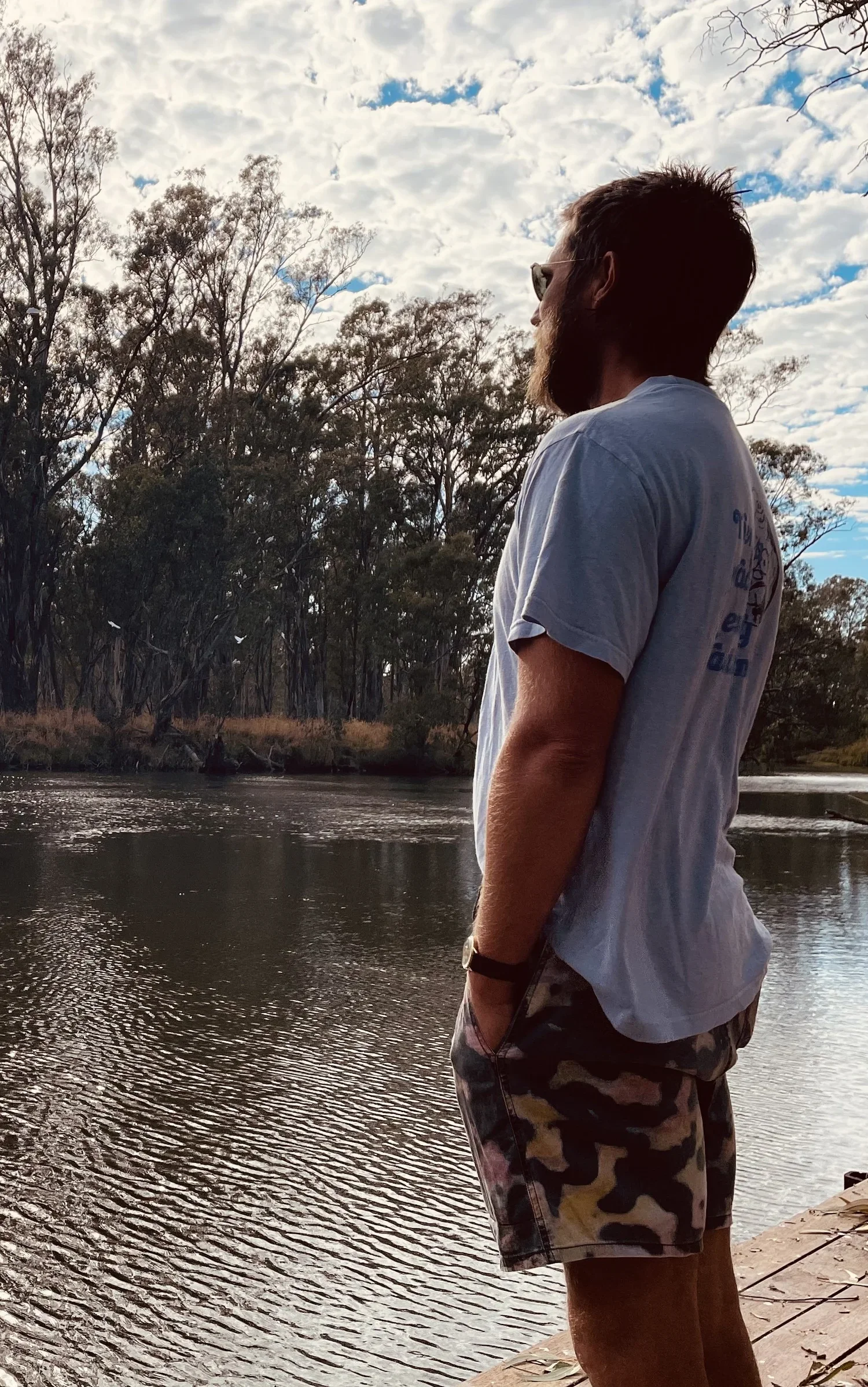 A man with a beard and sunglasses stands on a riverbank, looking at the water with trees and a cloudy sky in the background.