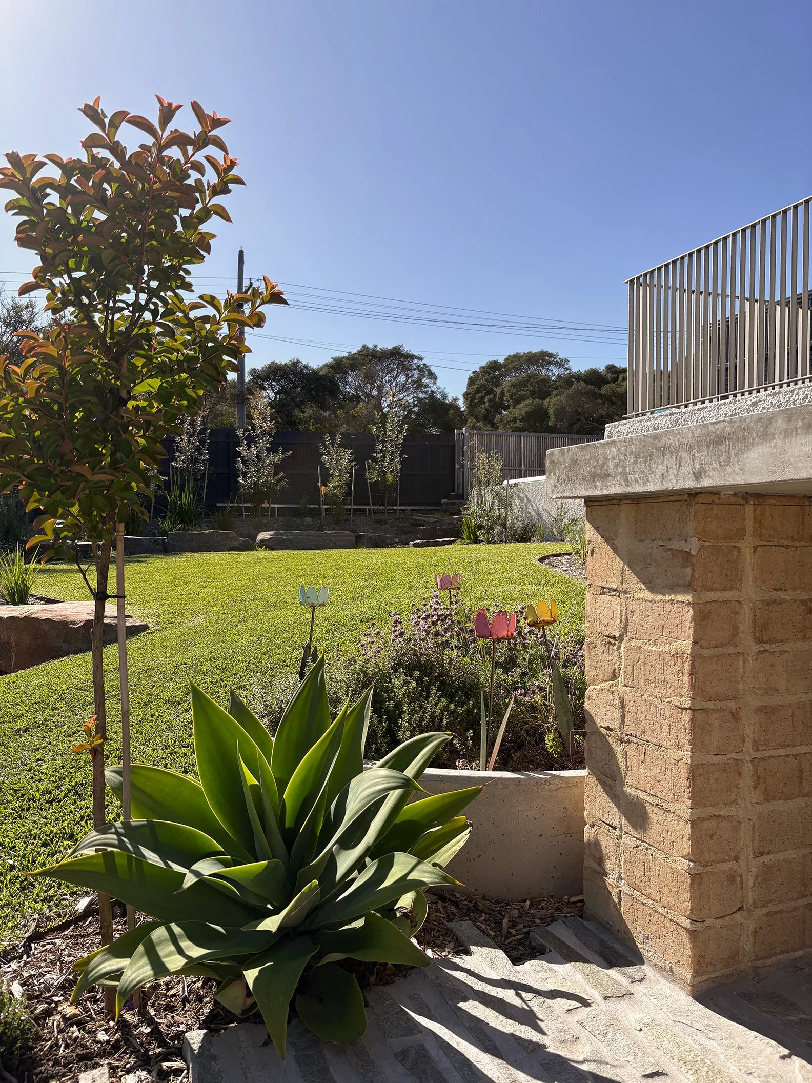 A sunny backyard garden with green grass, trees in the background, potted plants with colorful flower-shaped stakes, and a concrete brick column with a white railing on top.