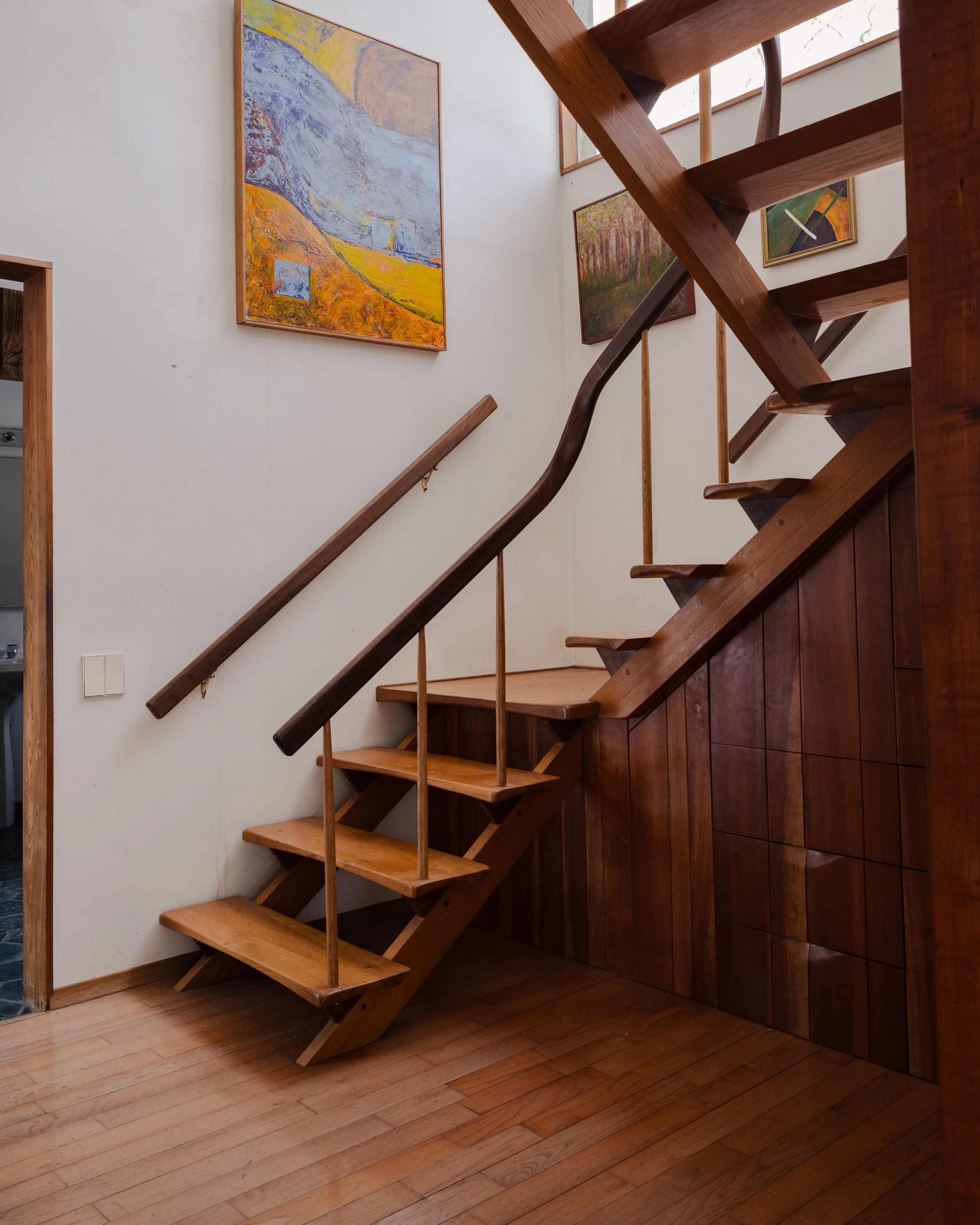 Editorial interior photograph of a modern wooden staircase featuring warm natural light, rich wood textures, minimalist railing design, and curated wall art in a contemporary residential space.