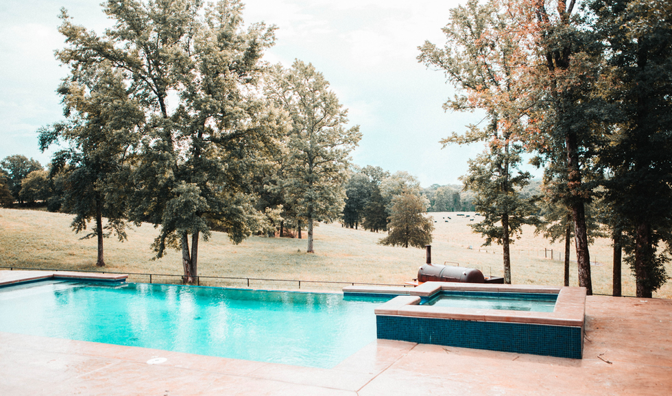 An outdoor swimming pool with a small attached hot tub, set against a grassy field with trees and a distant farm landscape.