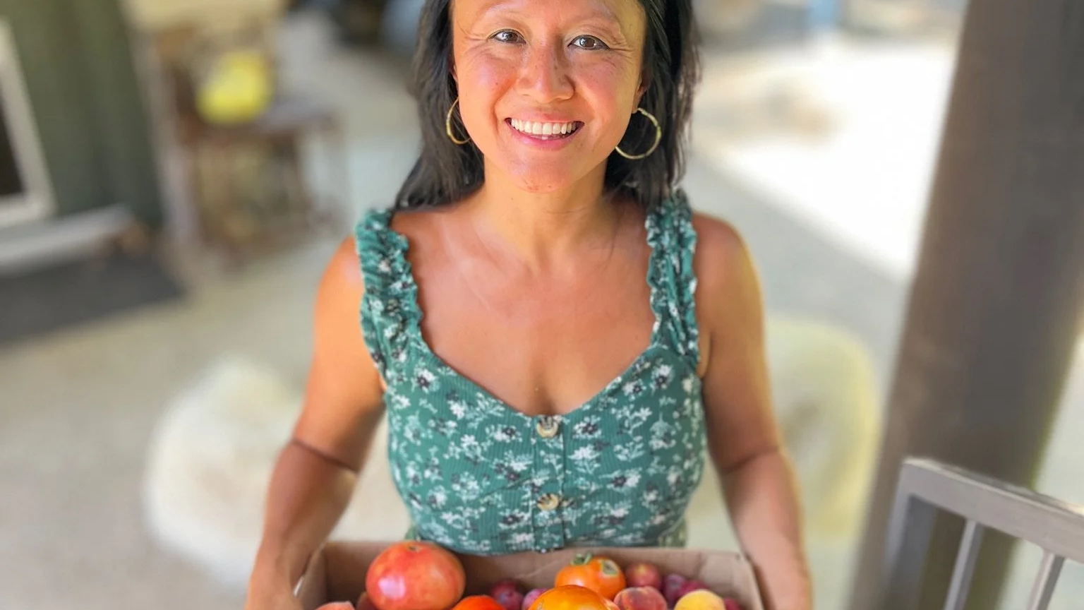A woman with dark hair, wearing a green floral dress and hoop earrings, smiling while holding a box of peaches and apples.