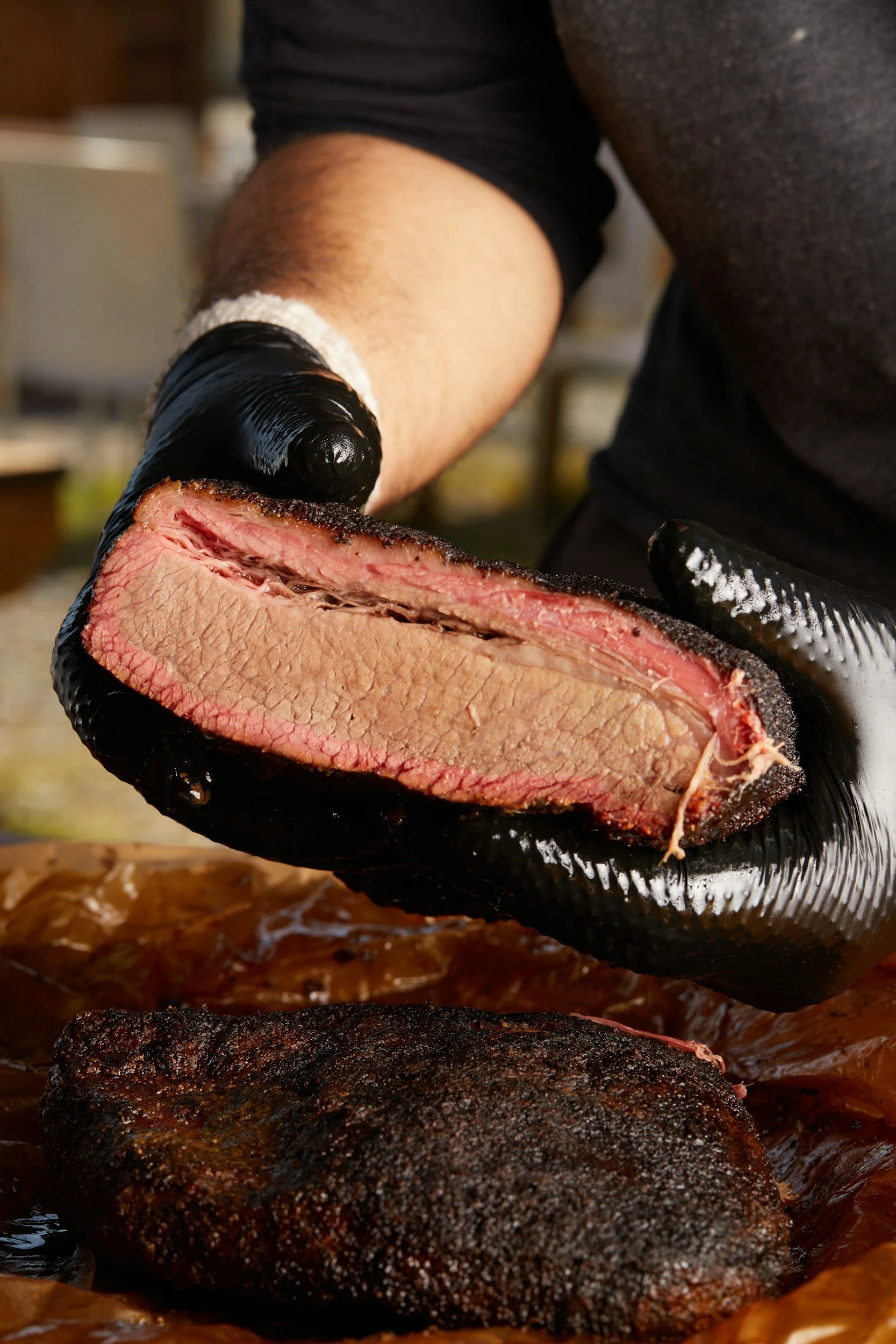 Person wearing black gloves holds a smoked brisket cut in half, showing the pink smoke ring and cooked interior, with another smoked piece of meat below on a wooden surface.