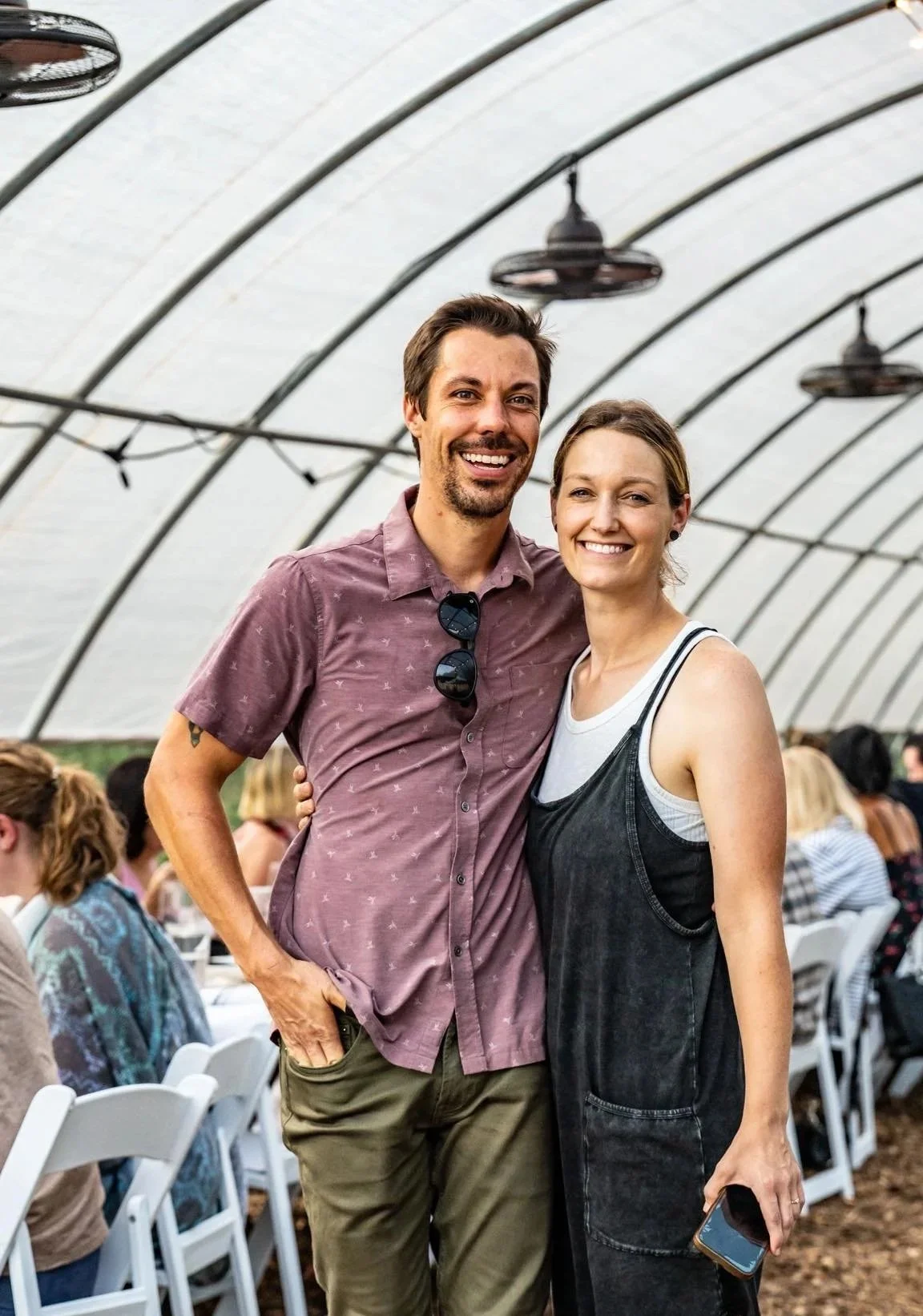 A smiling man and woman standing close together inside a greenhouse with arched ceiling, surrounded by seated people at tables, holding a smartphone.