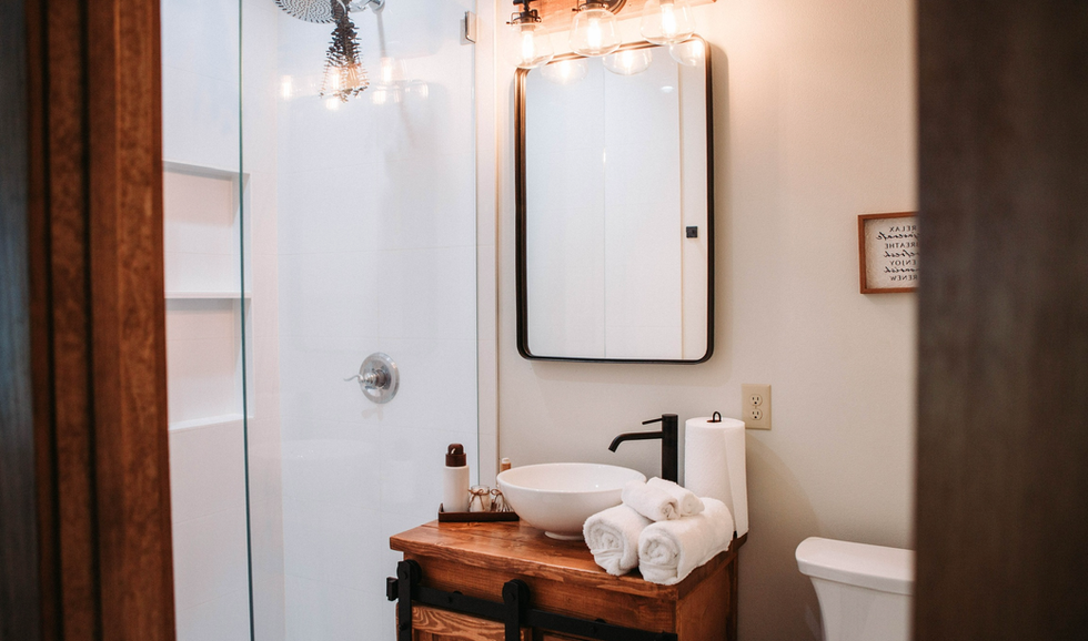 Bathroom with minimalist design featuring a wooden vanity, white vessel sink, black faucet, rolled white towels, mirror, shower area, and a small framed sign on the wall.