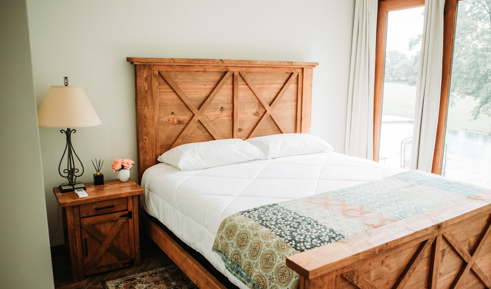A bedroom with a wooden bed frame, white bedding, and pillows, next to a nightstand with a lamp, flowers, and a small decorative item. There is a large window with curtains, showing an outdoor view.