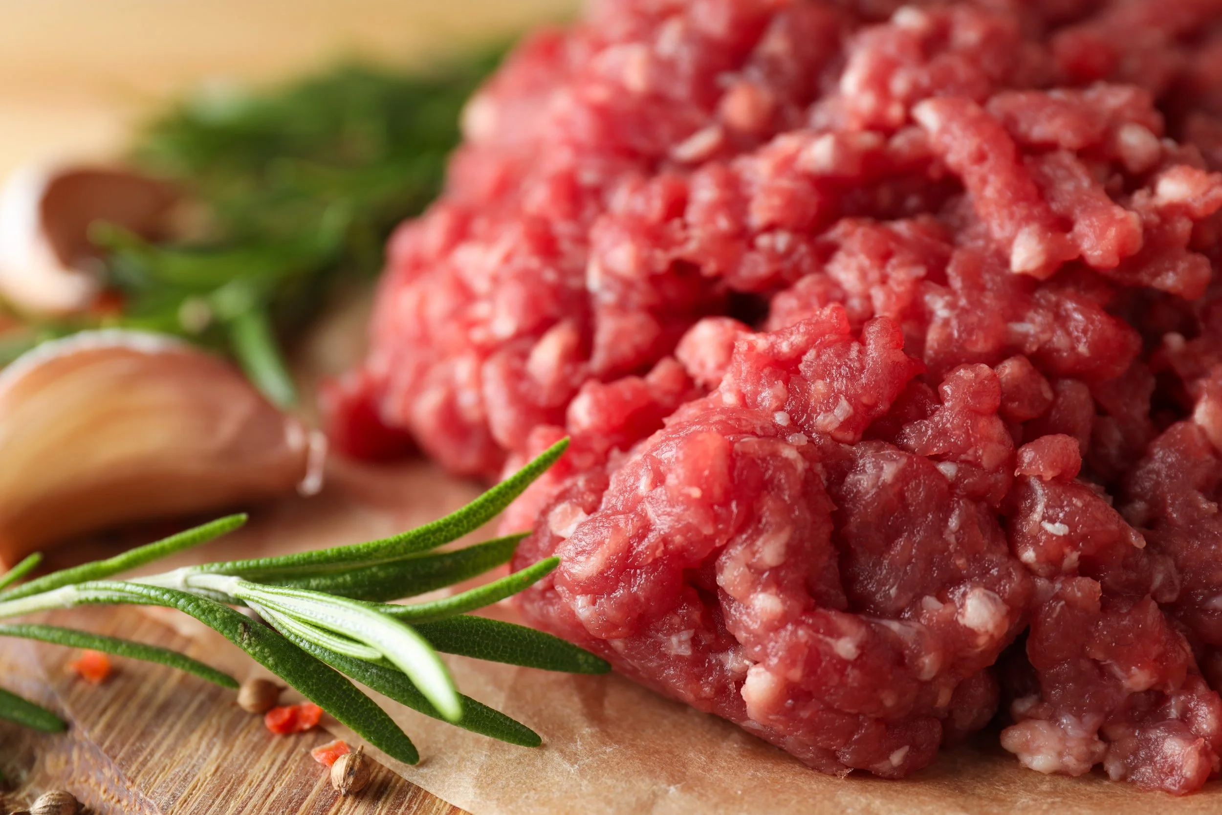Raw ground beef with garlic cloves, fresh rosemary, black peppercorns, and parsley on a wooden surface.
