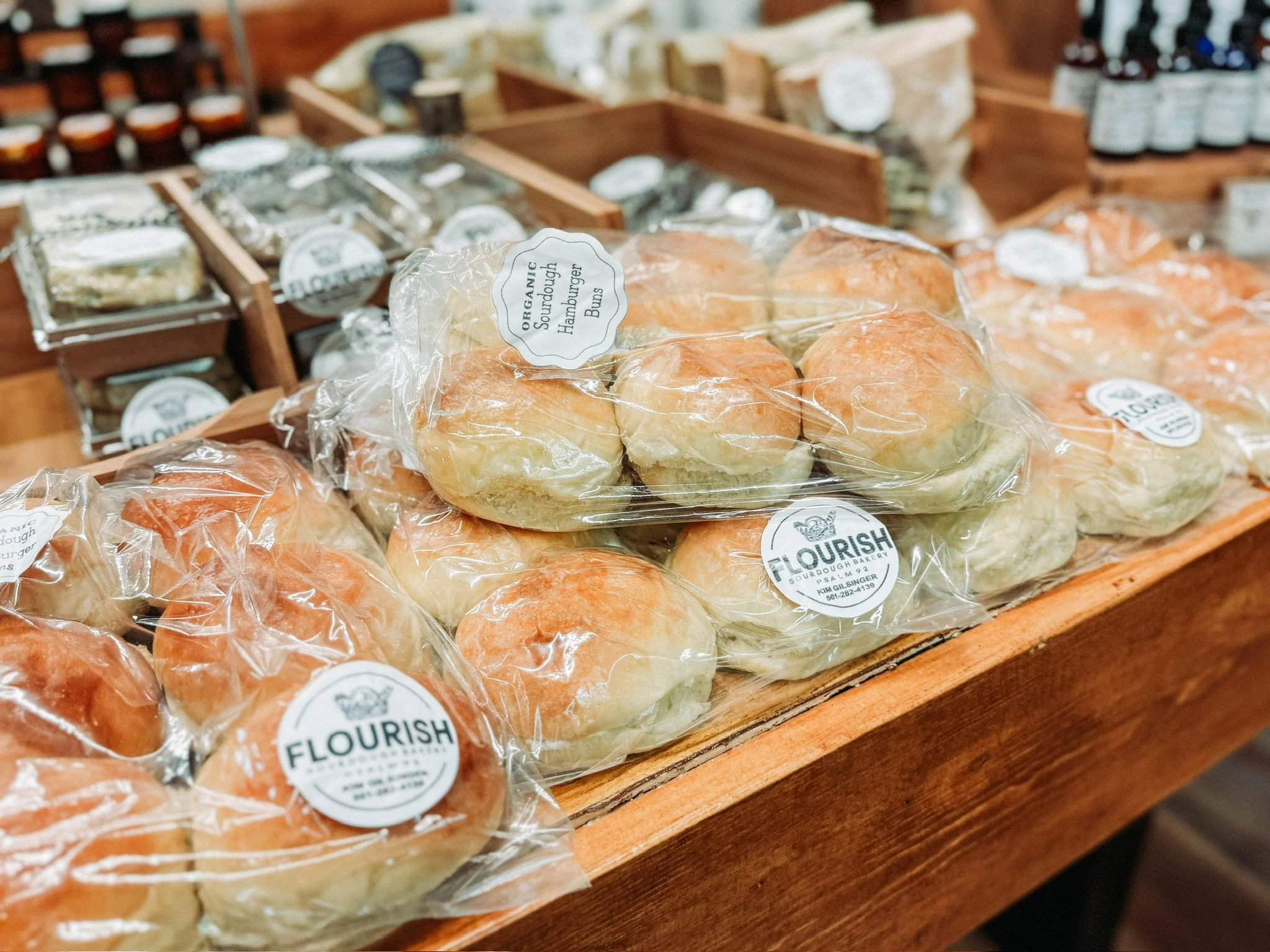 Bags of organic hamburger buns labeled 'Flourish' on a wooden display table in a bakery. In the background, there are other baked goods and jars.