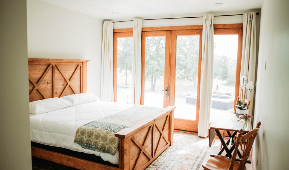 A bedroom with a wooden bed, white bedding, a small desk with a lamp and orchid, and large glass doors with white curtains overlooking a outdoor patio and green trees.