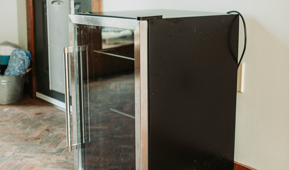 A small black and silver beverage refrigerator with a glass door, positioned against a beige wall.