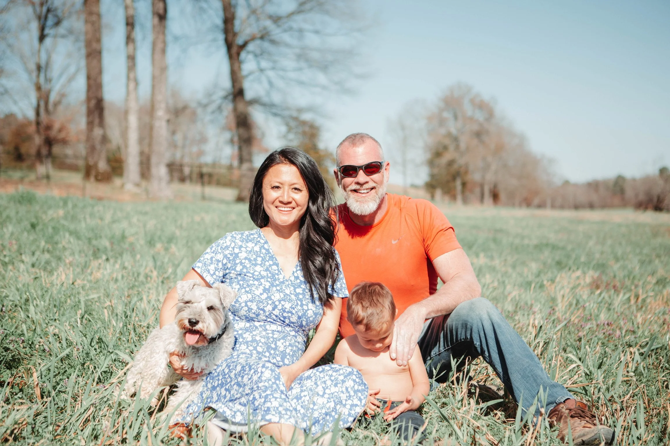 A happy family sitting on the grass in a park on a sunny day, with a woman, a man, a young boy, and a dog.