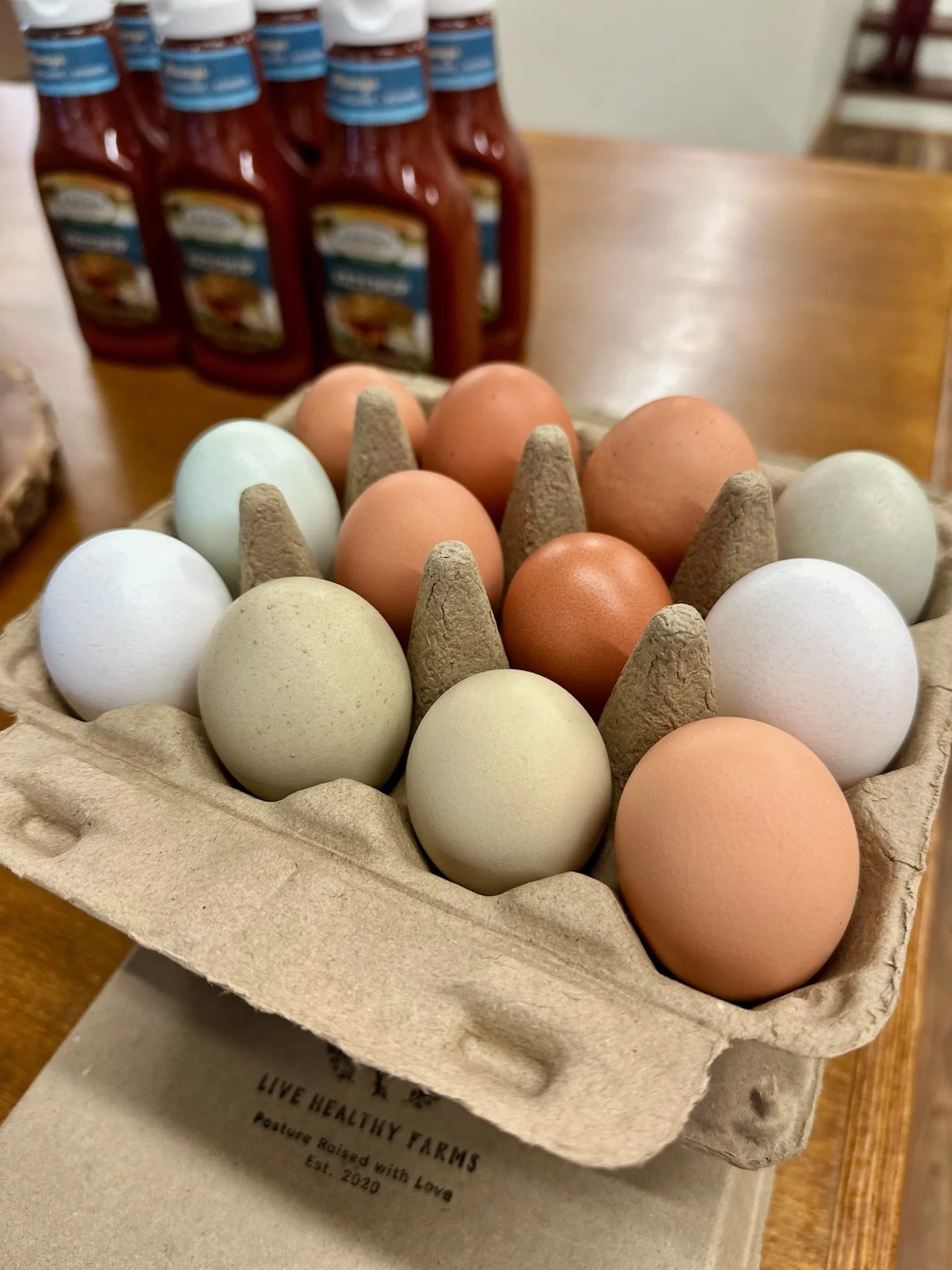 A carton of assorted eggs with different colors, some being white, light green, beige, and light brown, on a wooden table with bottles of ketchup in the background.