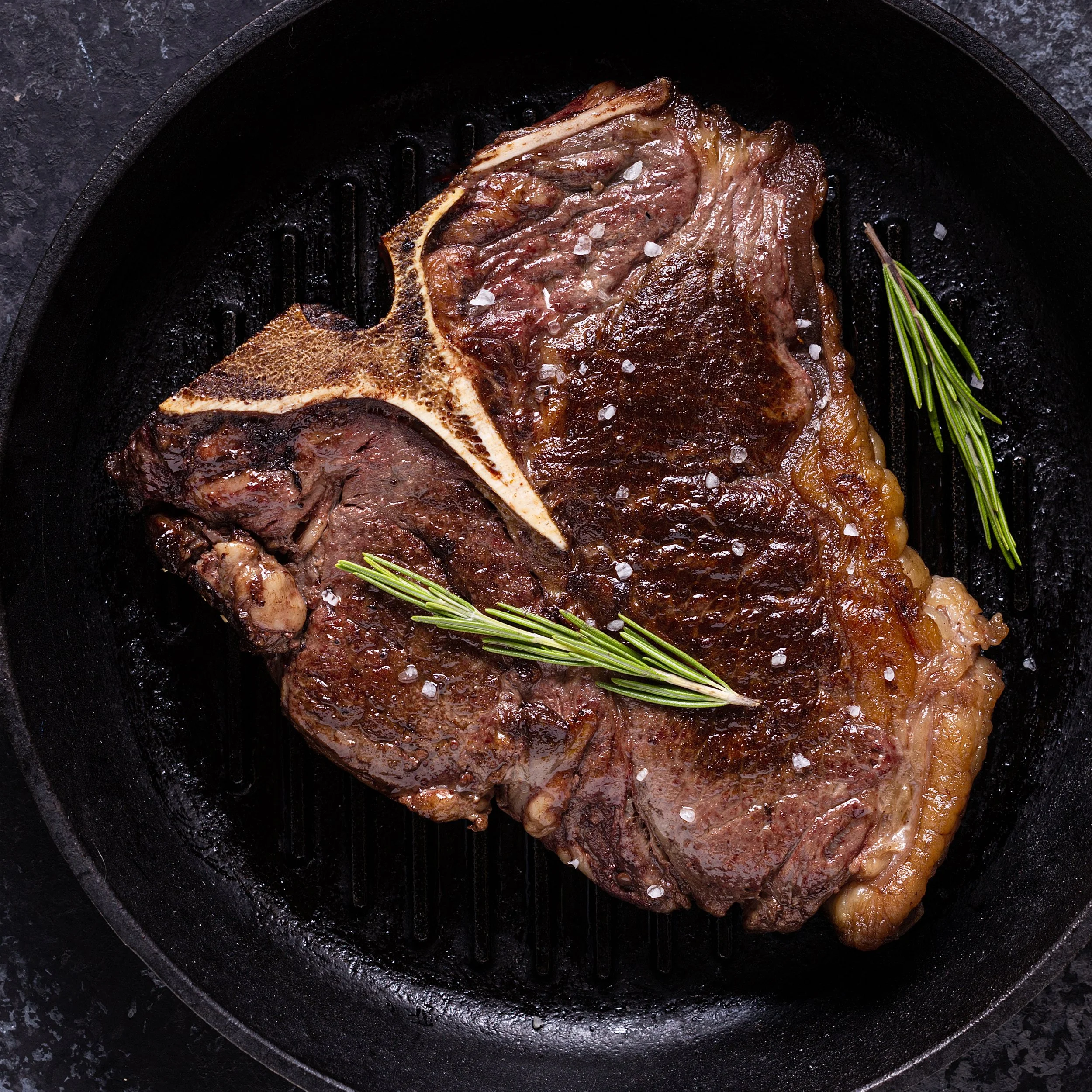 A cooked T-bone steak in a cast iron skillet garnished with rosemary sprigs and coarse salt.
