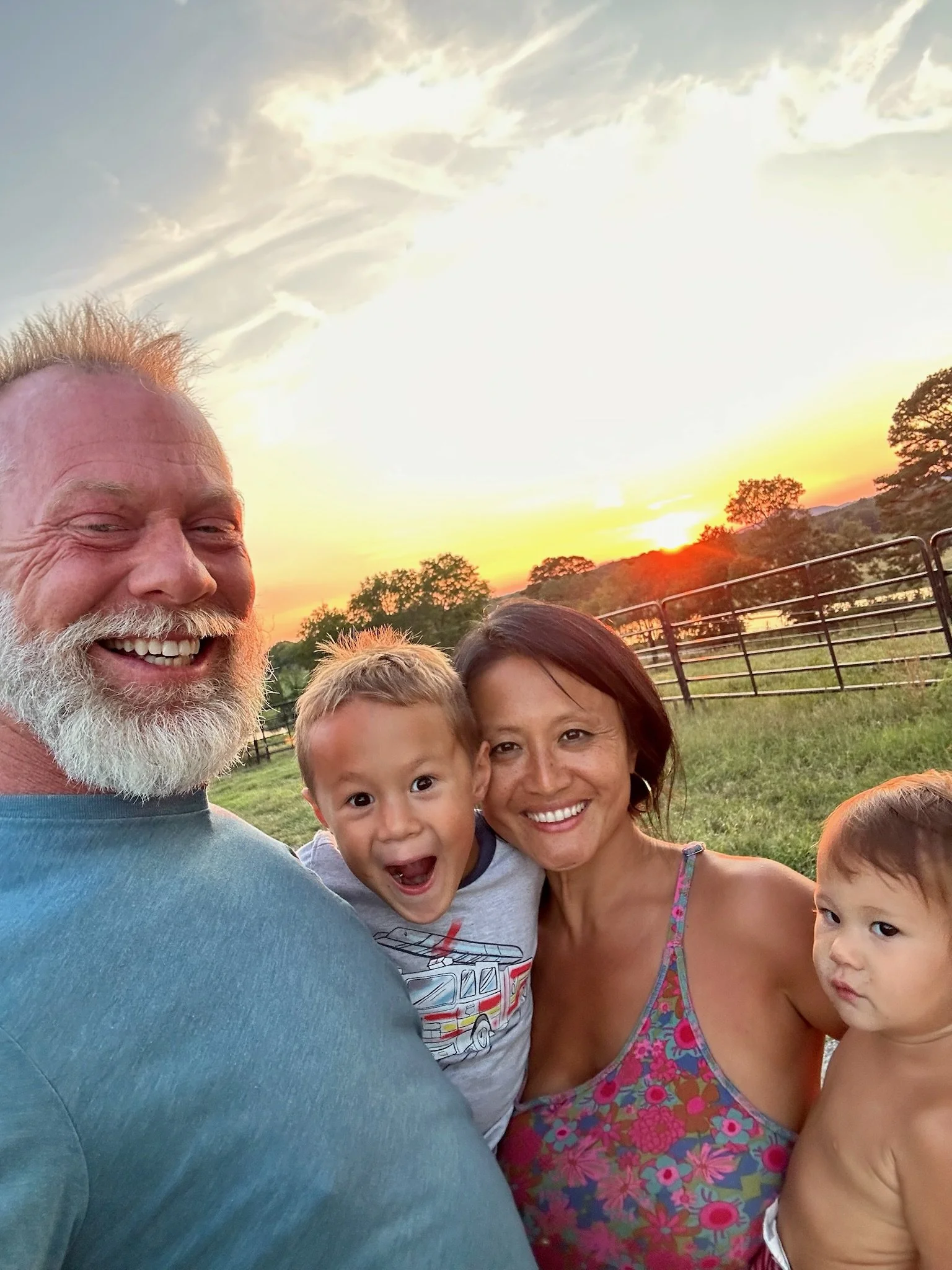 A family of four smiling and taking a selfie outdoors at sunset, with a fence and trees in the background.