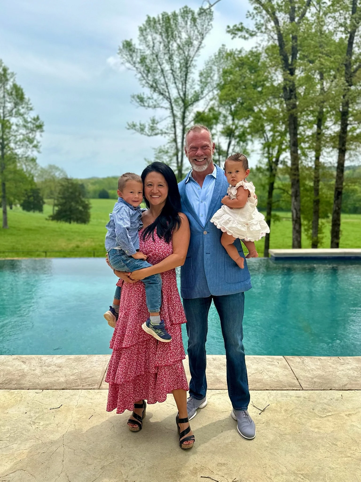 A family of four on a stone patio, with a swimming pool and green trees in the background. The woman is holding a boy in a blue shirt, and a man is holding a girl in a white dress. Everyone is smiling.