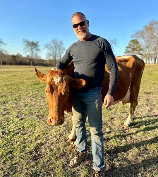 A man with sunglasses standing in a grassy field with a brown and white cow, holding the cow's head gently. Live Healthy Farms - Hot Springs, AR