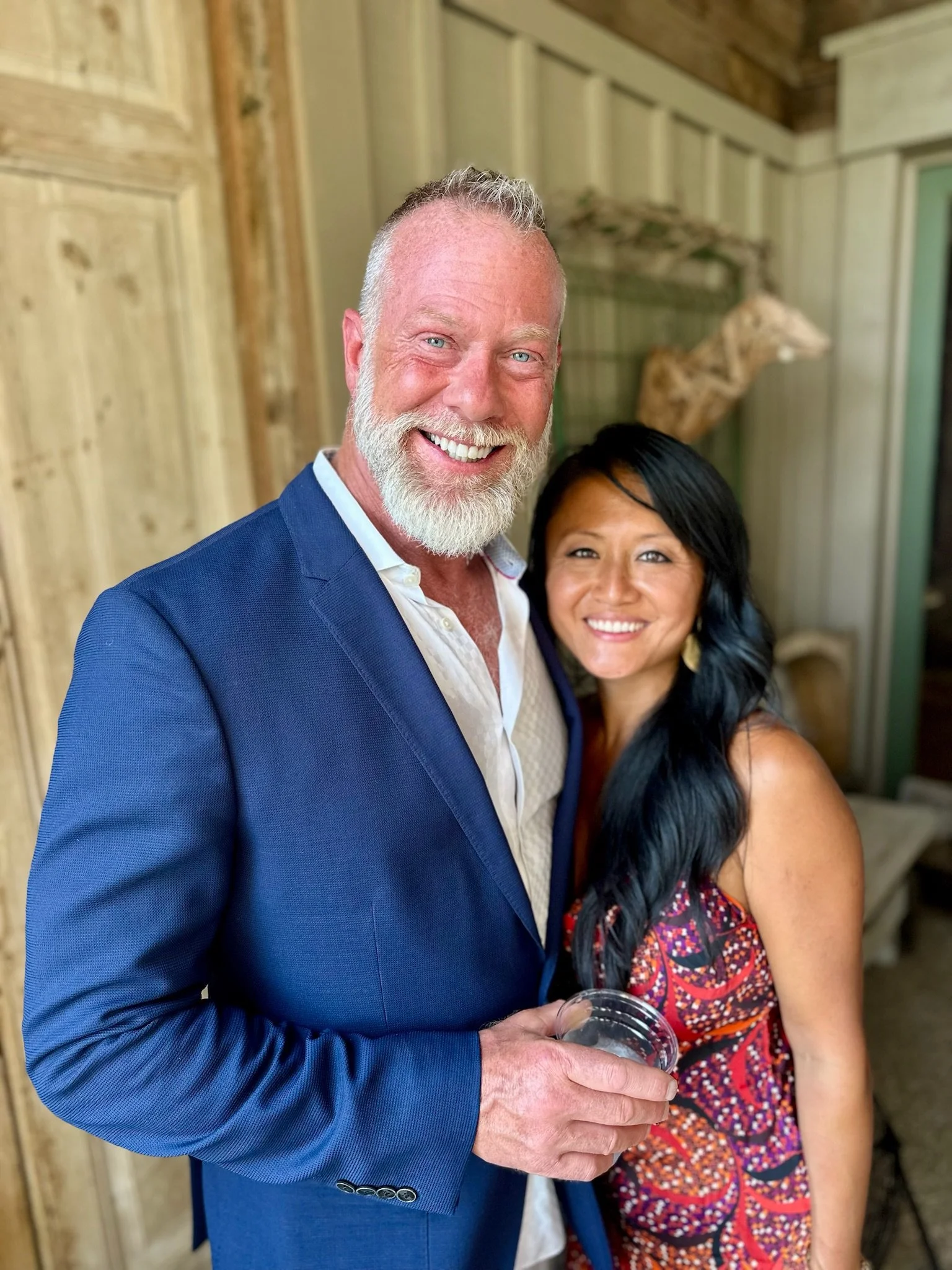 A smiling white man with a beard in a blue suit stands next to a smiling Asian woman with long black hair in a colorful dress. They are indoors with wooden walls and a carved wood decor in the background.