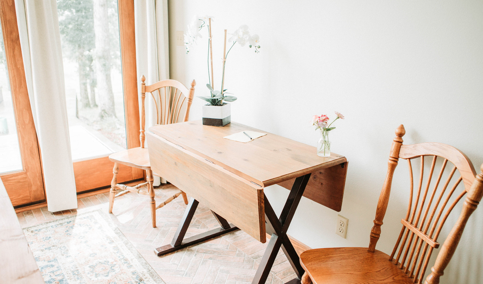 A wooden dining table with two wooden chairs, a vase with flowers, and a potted orchid near a window in a bright room.