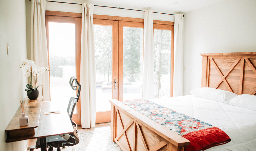 Bright bedroom with wooden bed, white bedding, quilt, and large glass doors with white curtains leading outside, near a wooden desk with a computer chair, a potted plant, and lamp.