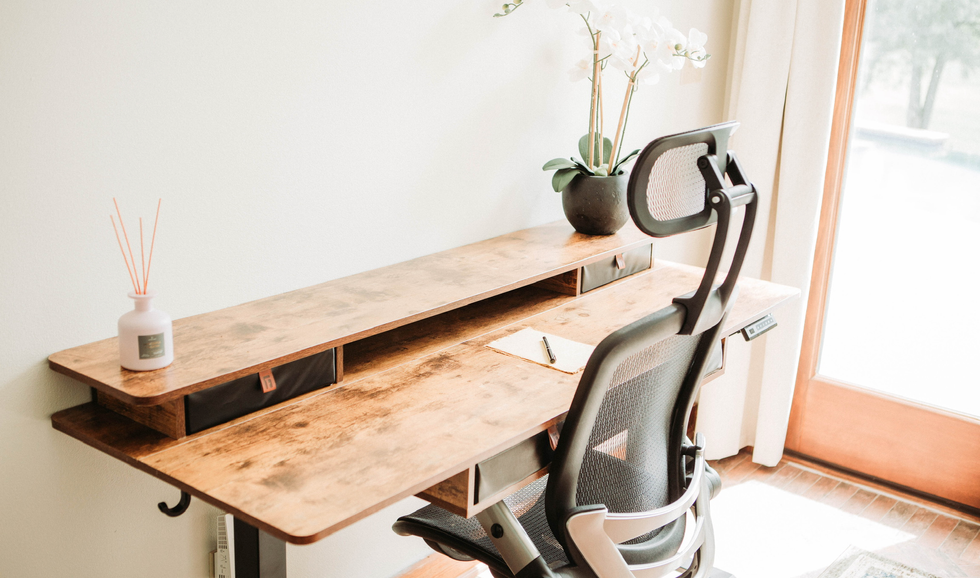 Home office desk with a black office chair, a potted orchid plant, a notepad with pen, and a diffuser or vase with reed sticks, near a glass door with curtains.