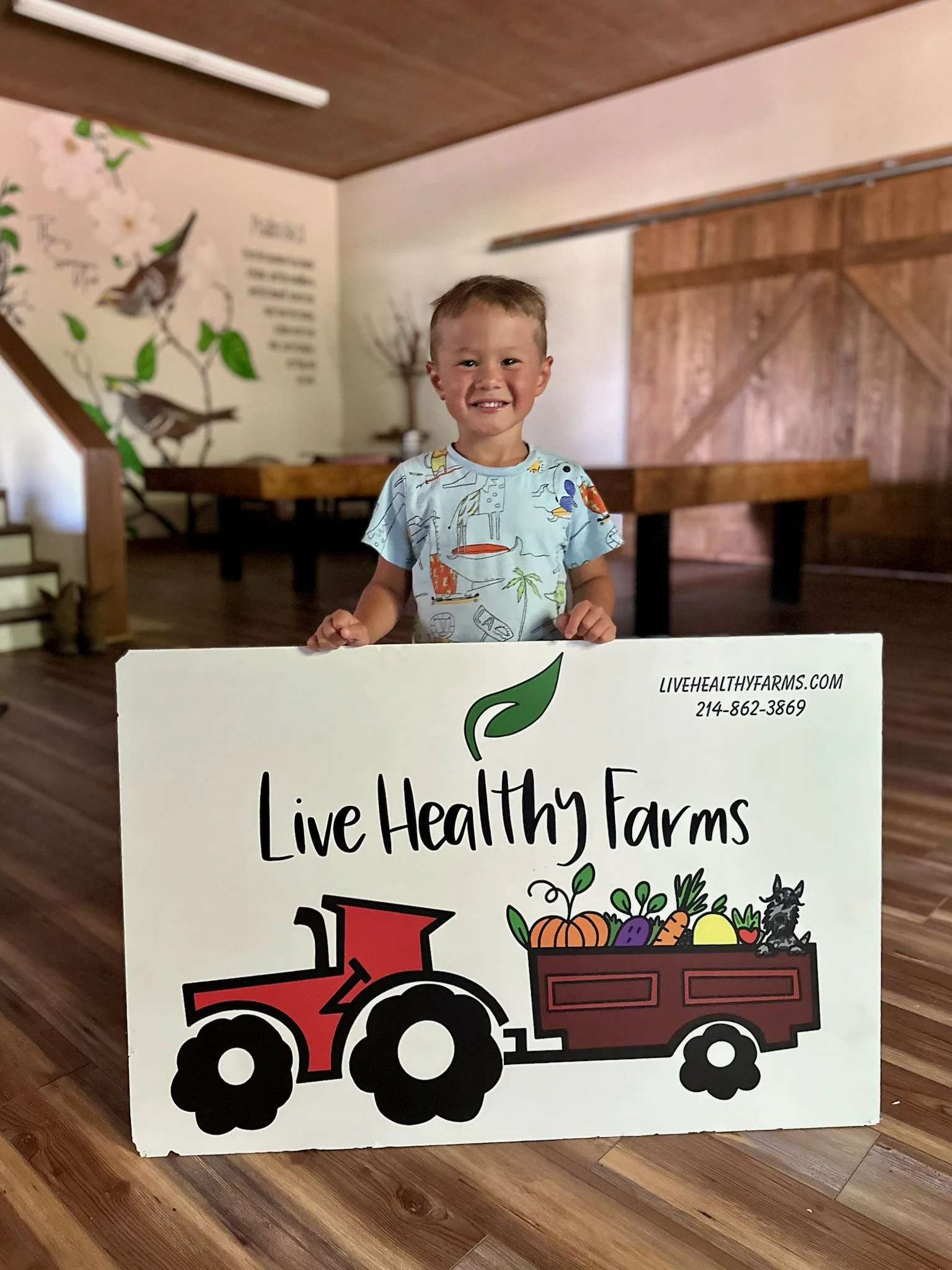A young boy with short hair smiling and holding a sign that reads "Live Healthy Farms" with a tractor and farm produce illustration. The background features wooden interior walls and a mural with birds and flowers.