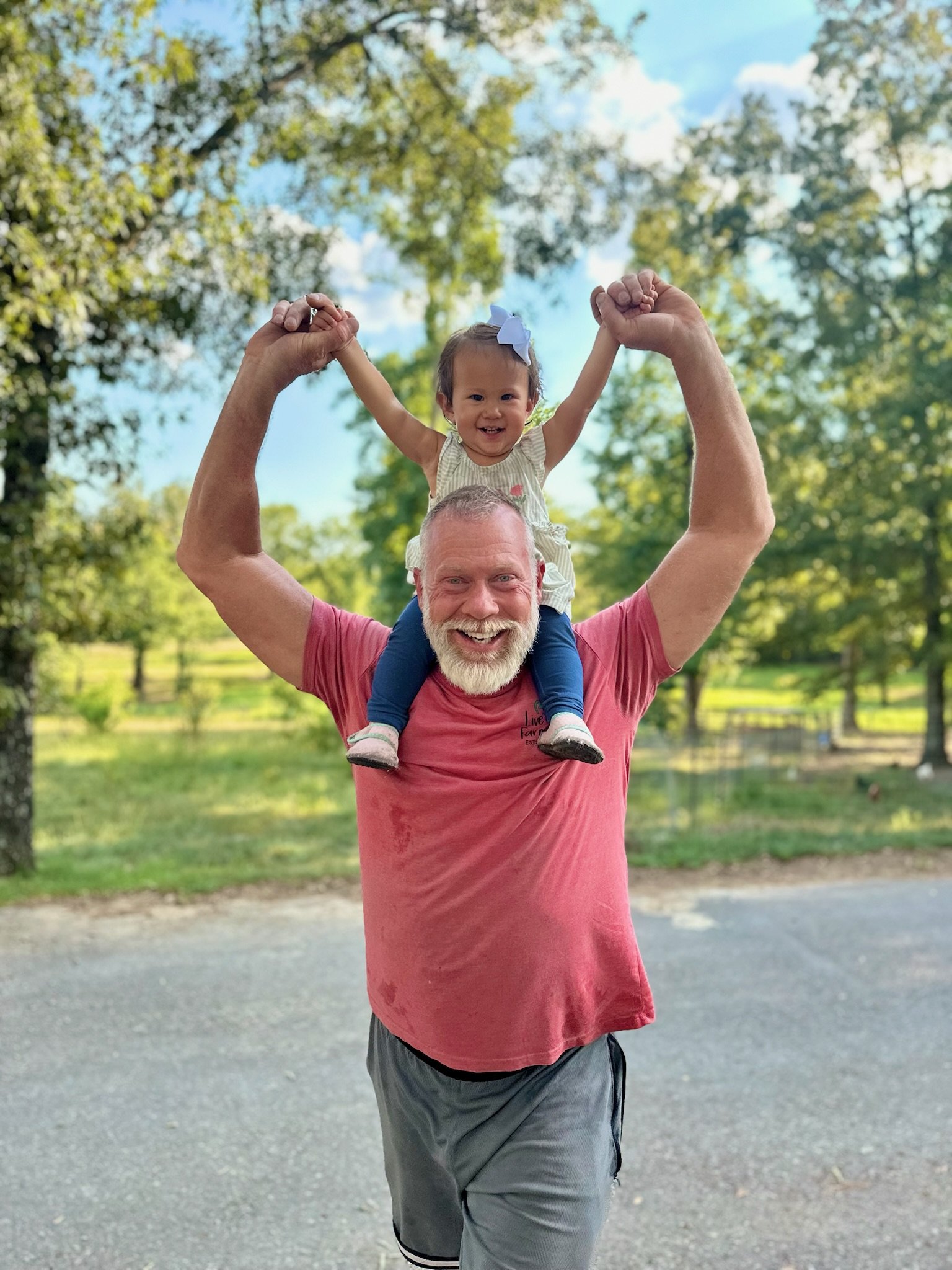 An elderly man with a beard and a young girl on his shoulders, both smiling, outdoors in a park or wooded area with trees and a gravel pathway in the background.