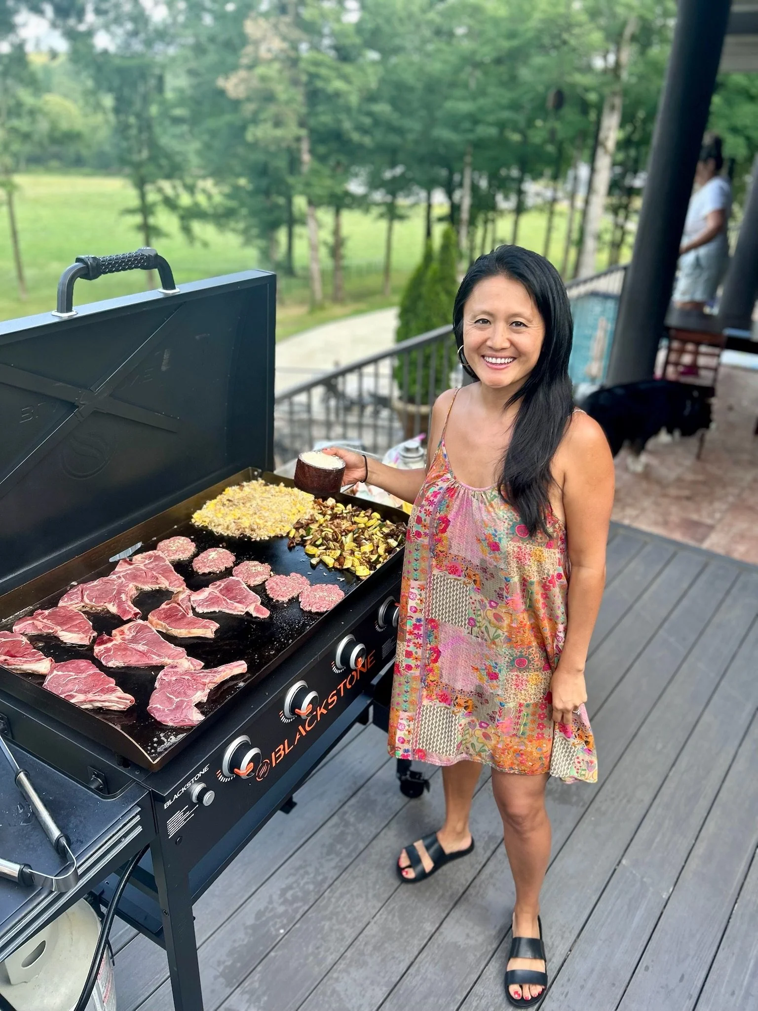 A woman with long dark hair and a colorful dress is smiling and standing next to a grill with raw meat and vegetables on it. She is holding a small bowl and standing on a wooden deck with a background of green trees and a grassy area.