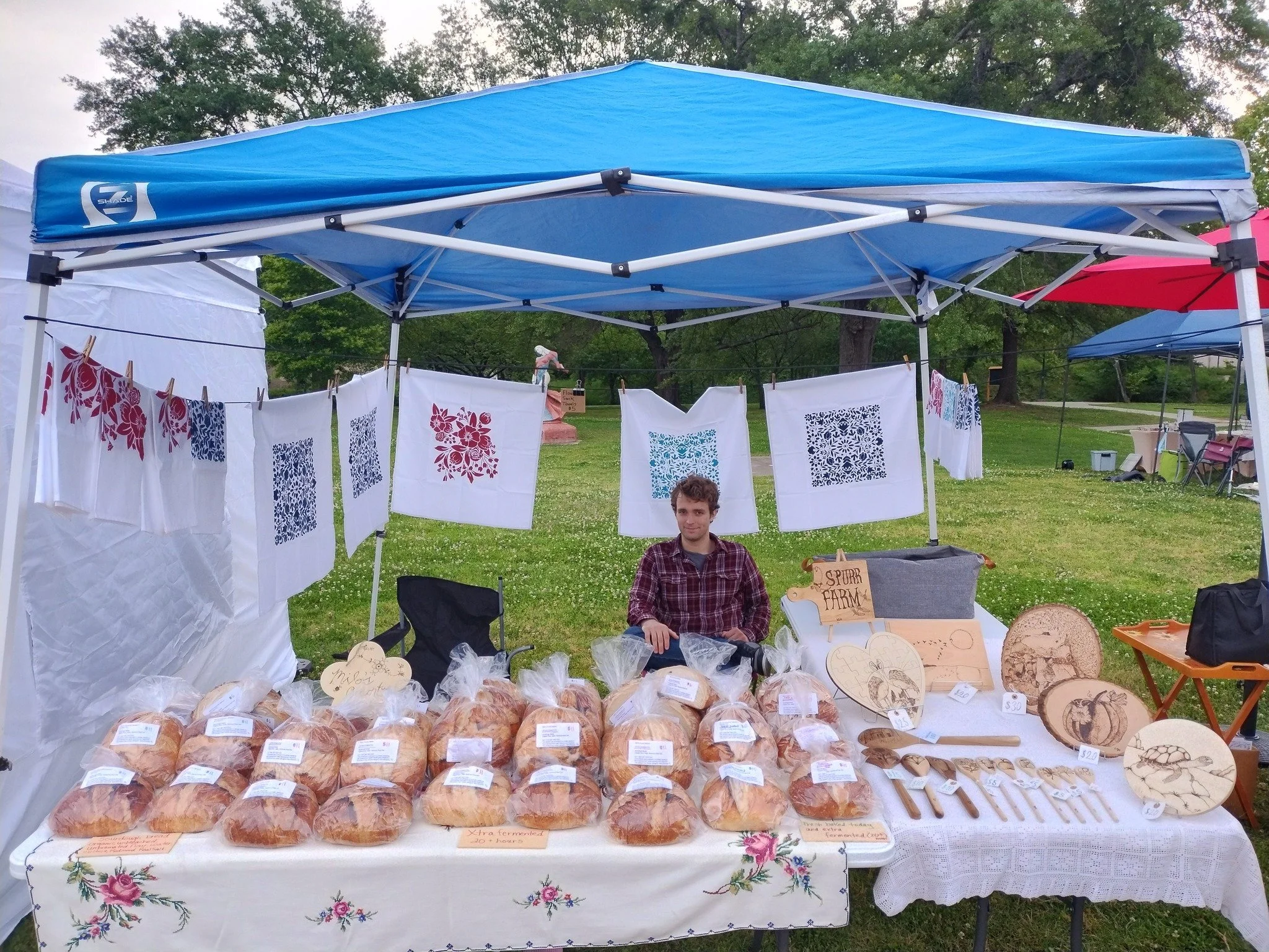 A vendor sitting at a craft stall under a blue canopy at an outdoor market, with various baked goods and handcrafted wooden items displayed on a table in front of him.