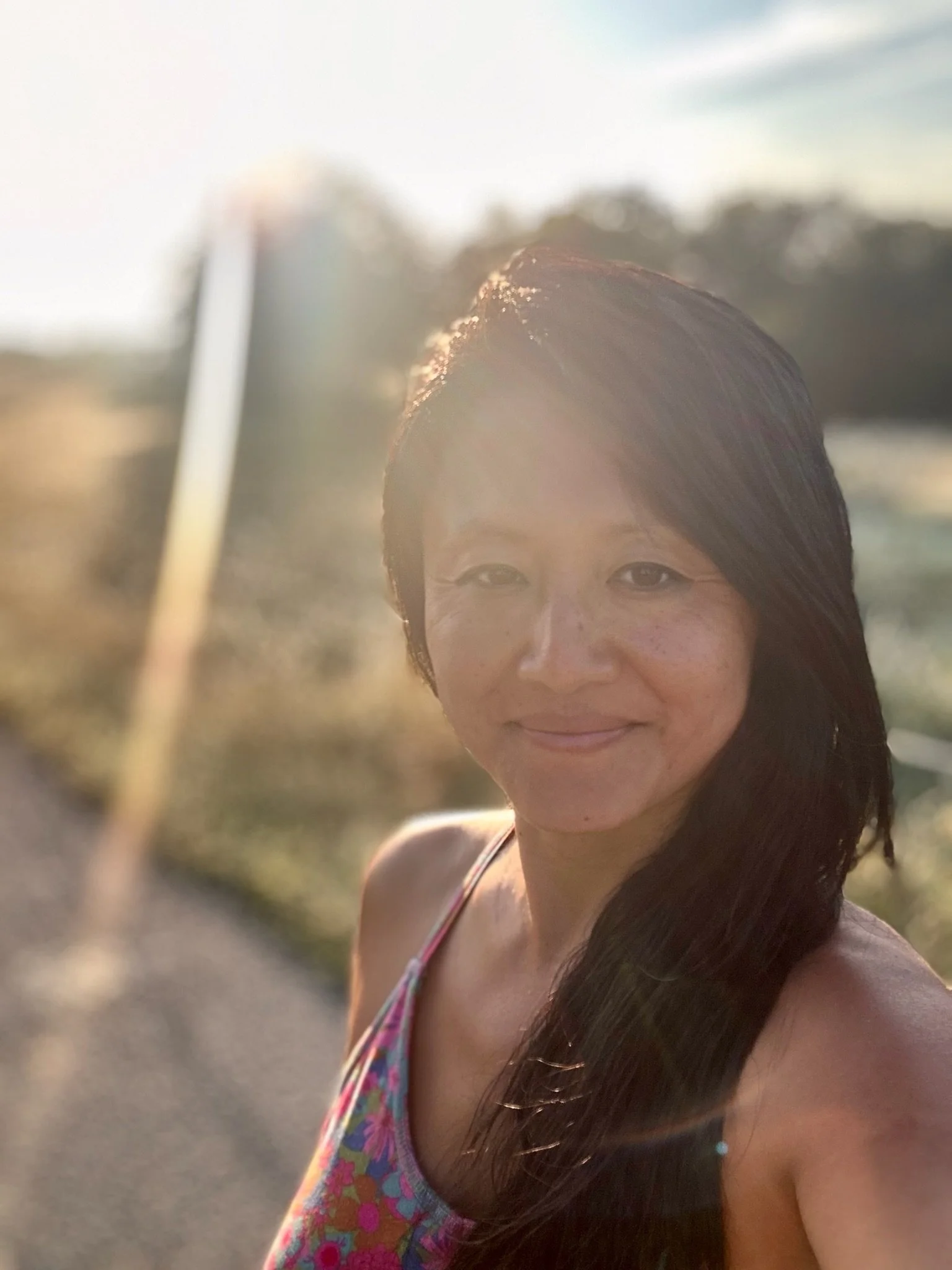A woman with long dark hair and freckles smiling outdoors at sunset, wearing a colorful tank top.