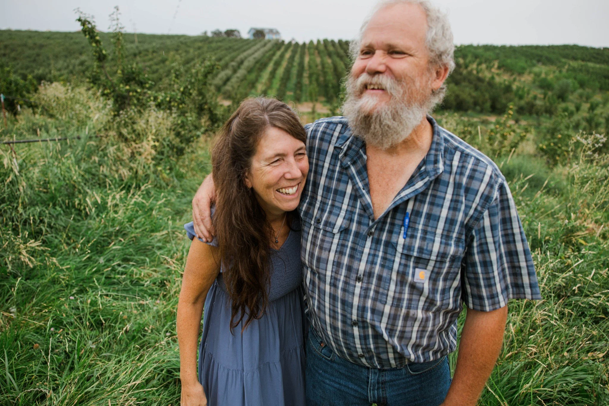A woman and an elderly man smiling and hugging outdoors in a grassy area with farmland and rows of trees in the background.
