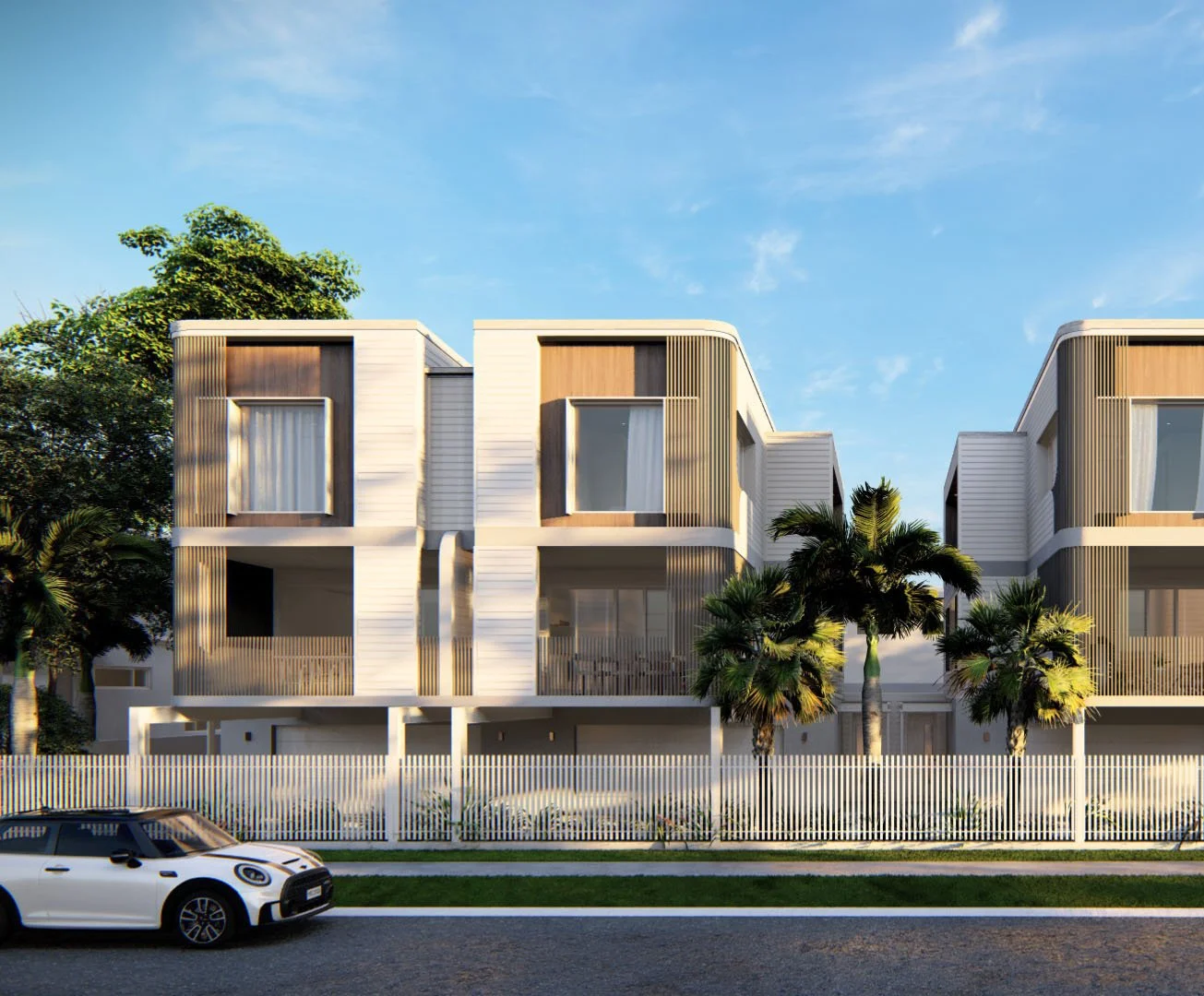 Modern multi-story residential buildings with white and wooden exterior, surrounded by trees and white fence, parked white car in front, under a blue sky.